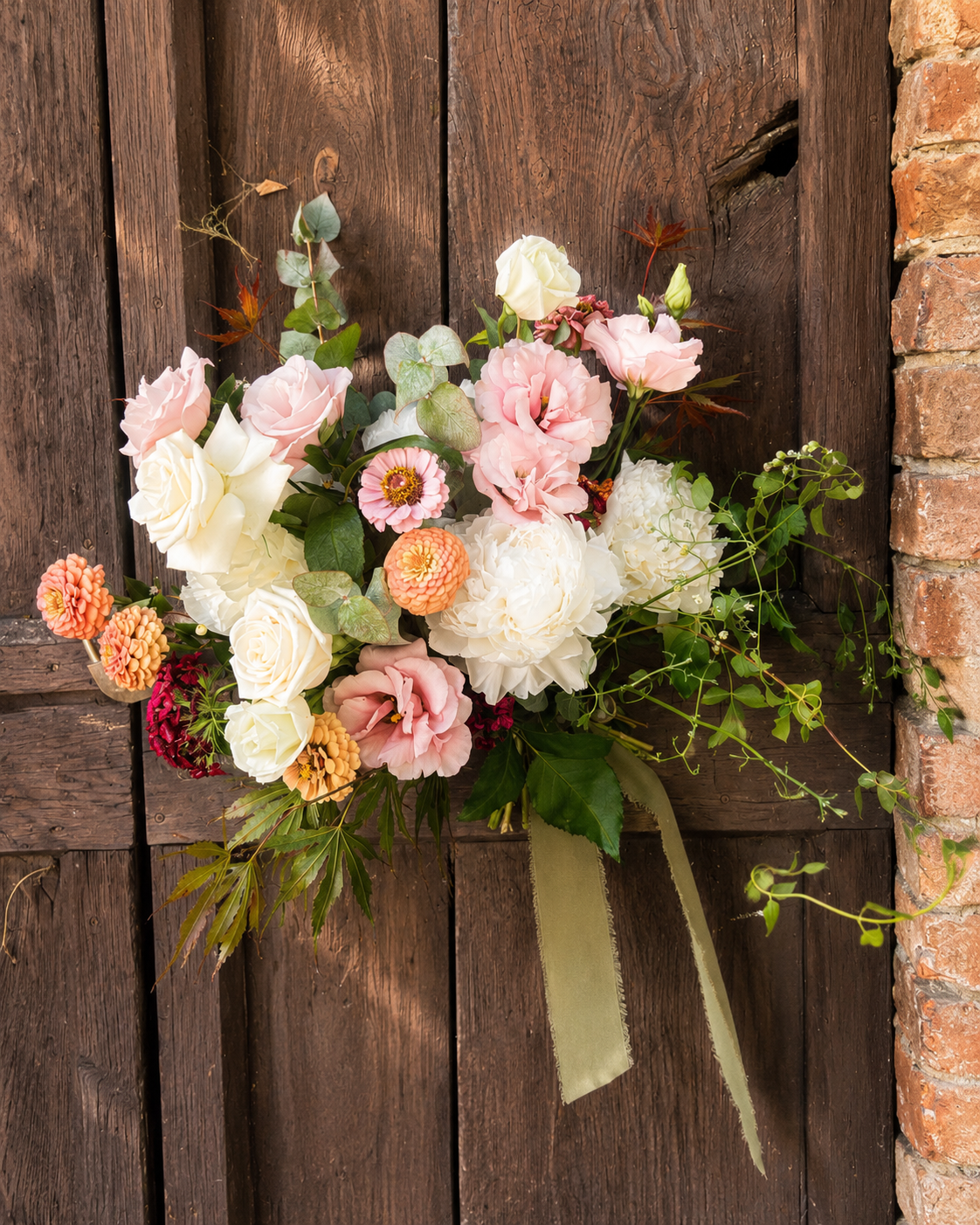A floral arrangement with white, pink, and peach flowers, surrounded by green leaves, attached to a rustic wooden door with a green ribbon.