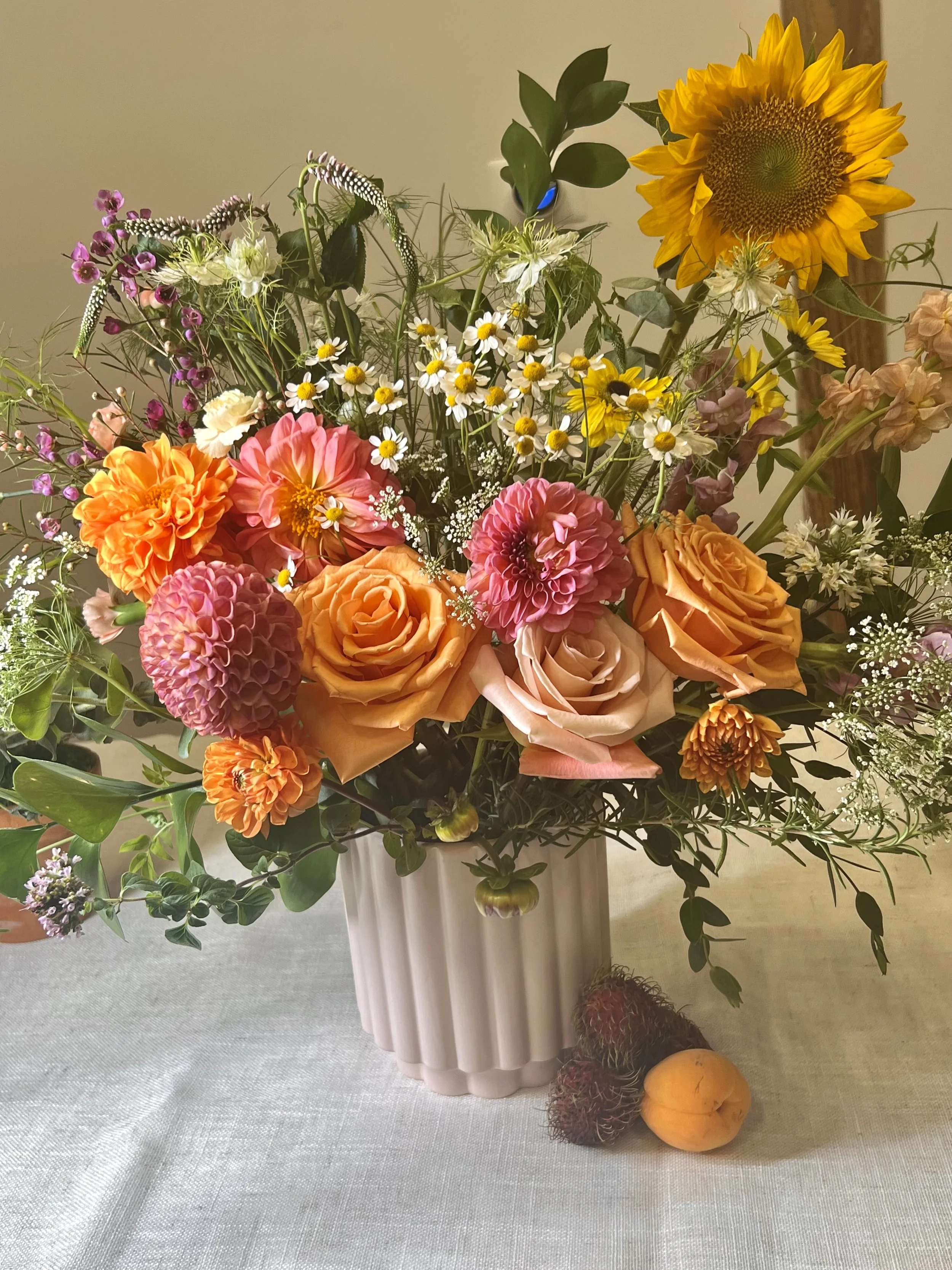 A bouquet of colorful flowers including sunflowers, roses, daisies, and dahlias in a white ceramic vase, placed on a table with some tropical fruits nearby.