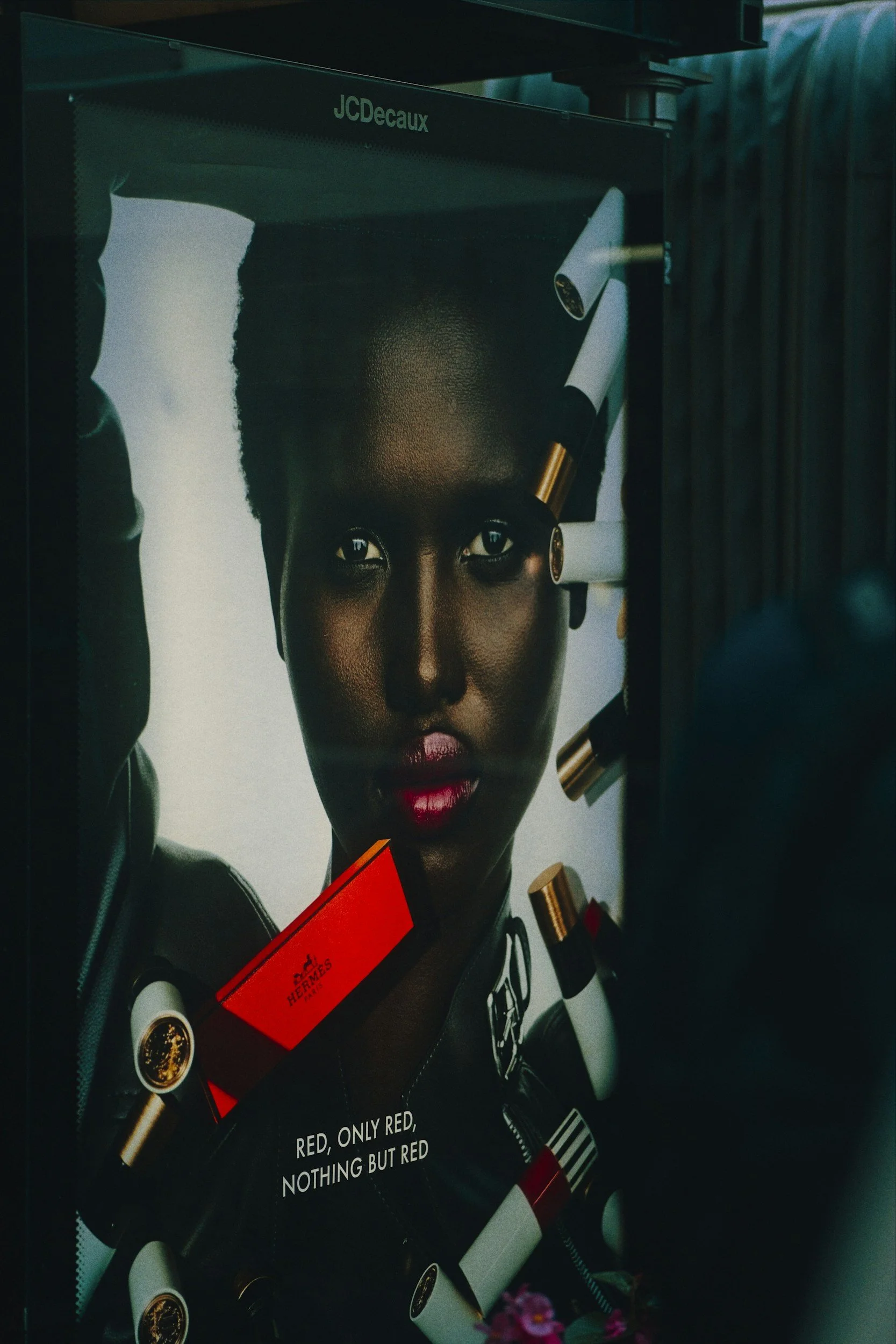 A poster of a woman with dark skin, short hair, and red lipstick, wearing a black leather jacket with patches, with an orange Hermès product, and the text "Red, Only Red, Nothing But Red." The poster is displayed in a dark setting with some pink flowers at the bottom.