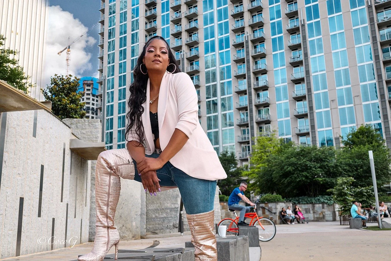 A woman with long black hair, hoop earrings, and a white blazer poses confidently in an urban park with a cityscape and high-rise buildings in the background. She is wearing high-heeled, patterned boots, dark jeans, and a black top.