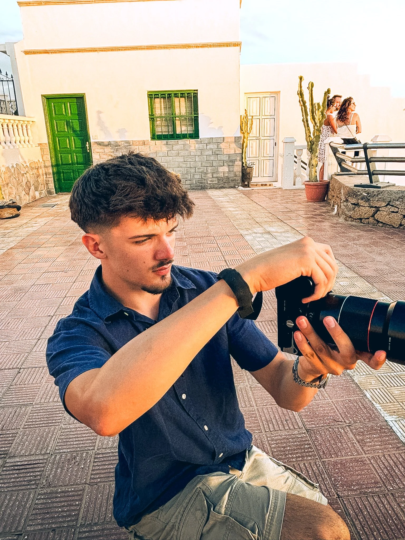 Young man with dark hair and facial hair, wearing a dark blue shirt and khaki shorts, taking a photo with a professional camera on an outdoor terrace during daylight, with two women standing near a tall cactus and white wall in the background.