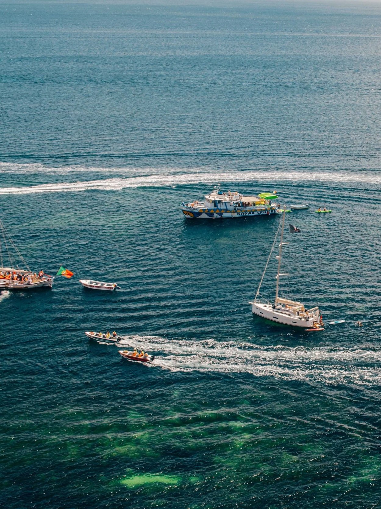 Multiple boats, including a large colorful kayak cruise boat and various smaller boats, sailing on the ocean with visible wake trails and green algae or seaweed on the water surface.