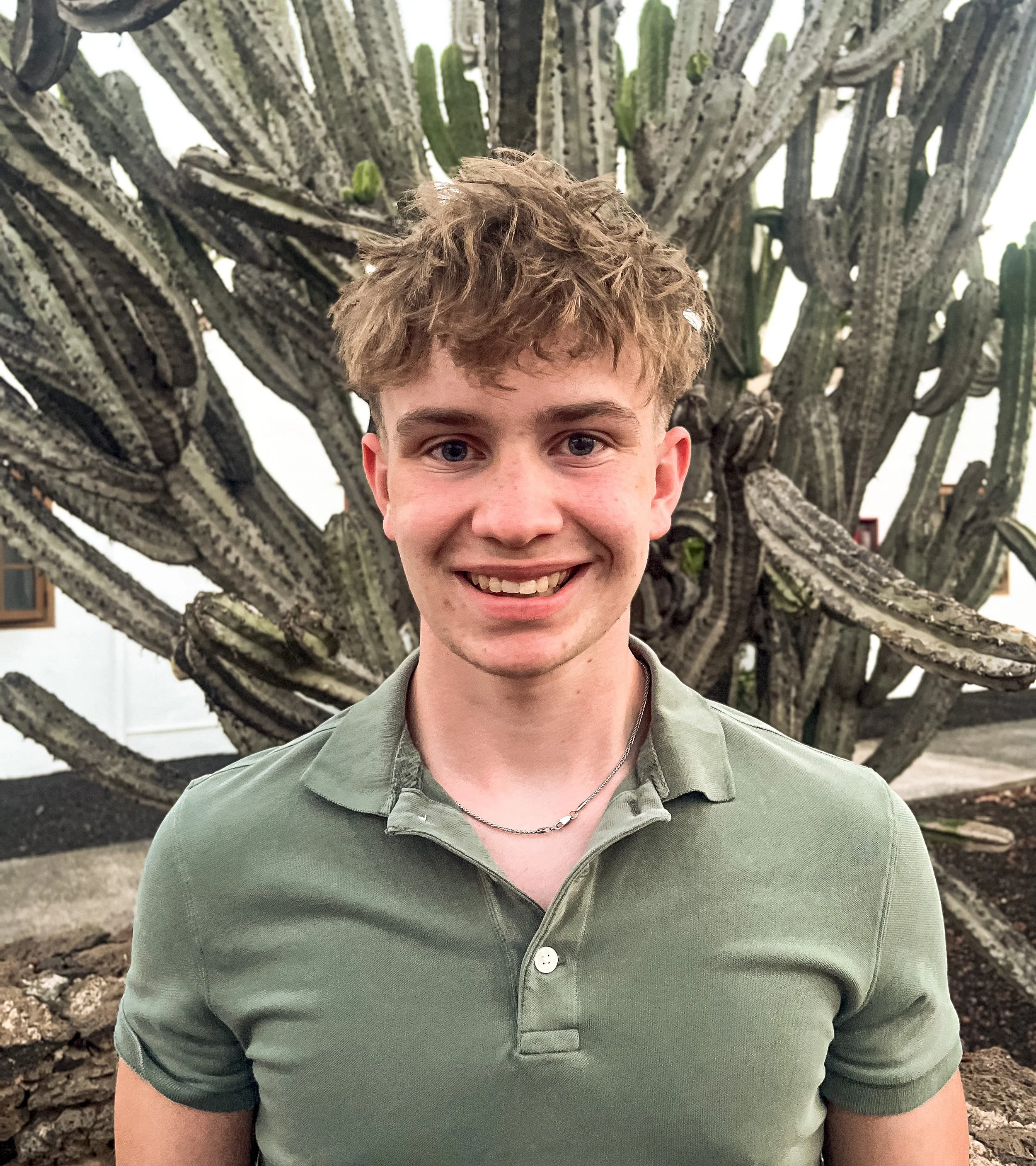 A young man with brown curly hair smiling, wearing a green button-up shirt, standing in front of a large cactus with multiple arms and spines.