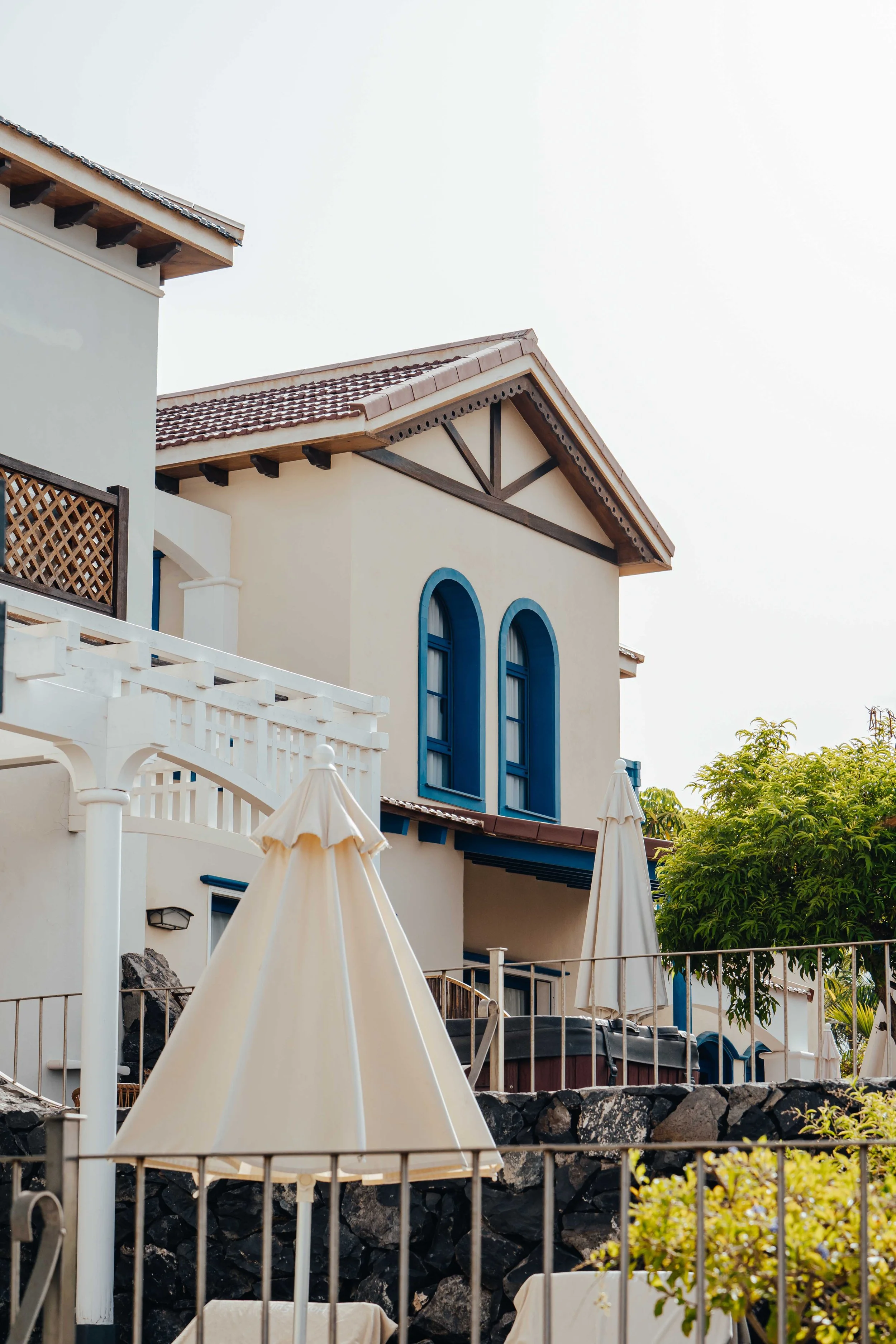 A multi-story building with a Mediterranean style, featuring arched windows with blue trim, a gabled roof with wooden support beams, and a white balcony with a lattice railing. There are umbrellas and outdoor furniture in the foreground, and a lush tree to the right.