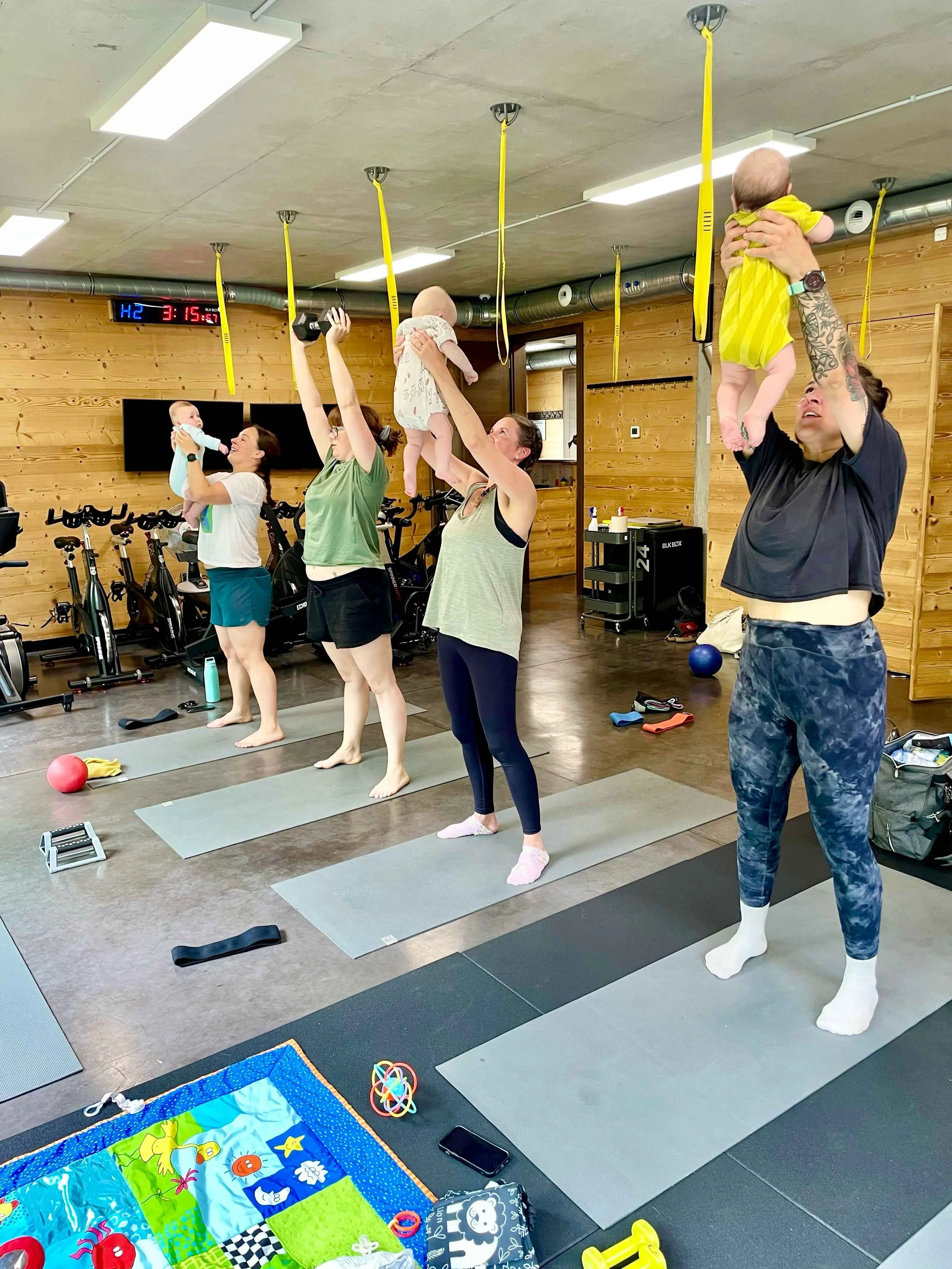 A group of women and two babies participating in a baby workout or fitness class in a gym with wooden wall paneling. Some women are lifting babies in the air or holding them, and exercise mats and toys are visible on the floor.
