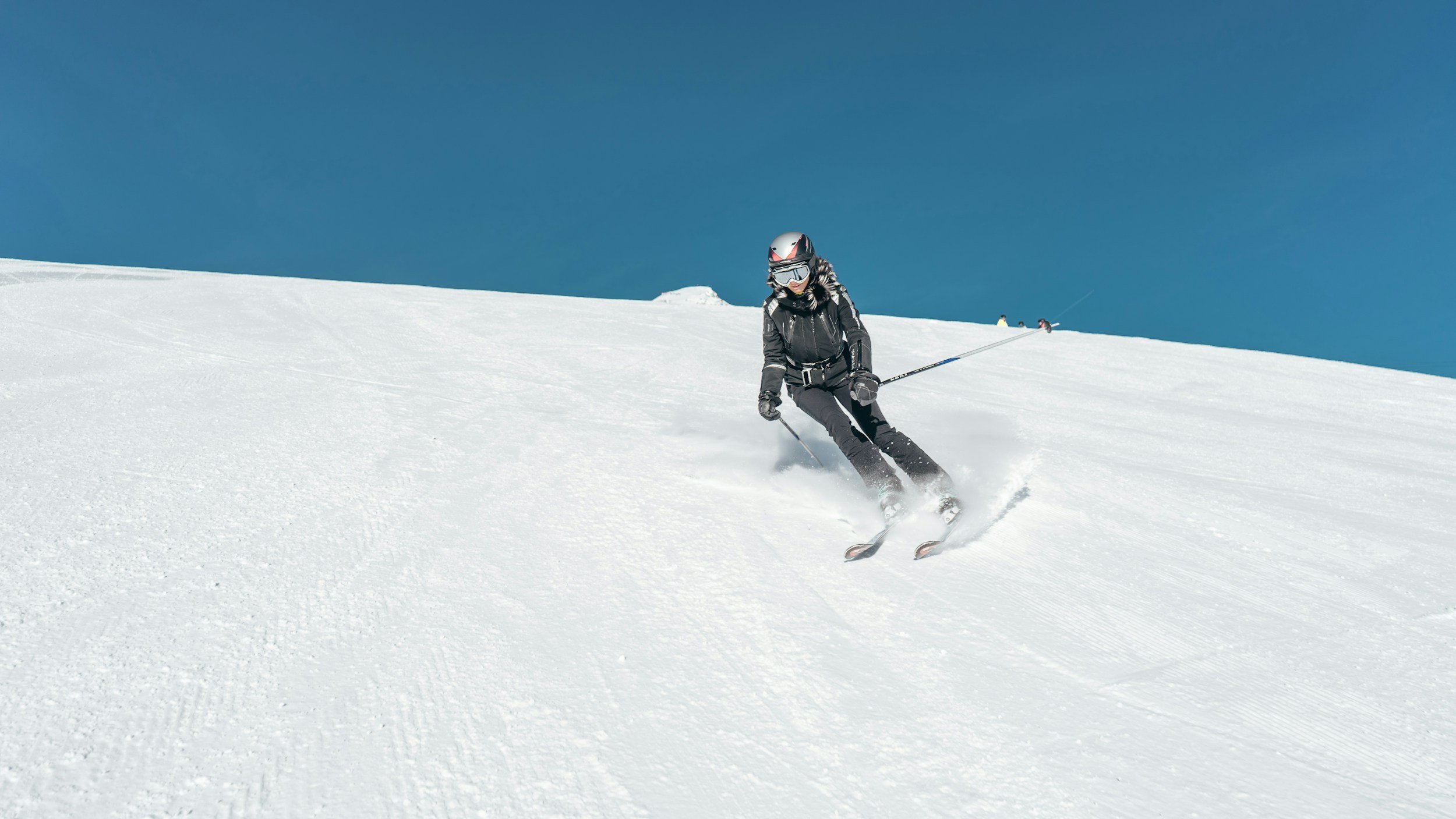 A person skiing down a snow-covered slope on a clear, sunny day, wearing a black jacket, black pants, a helmet, and goggles.