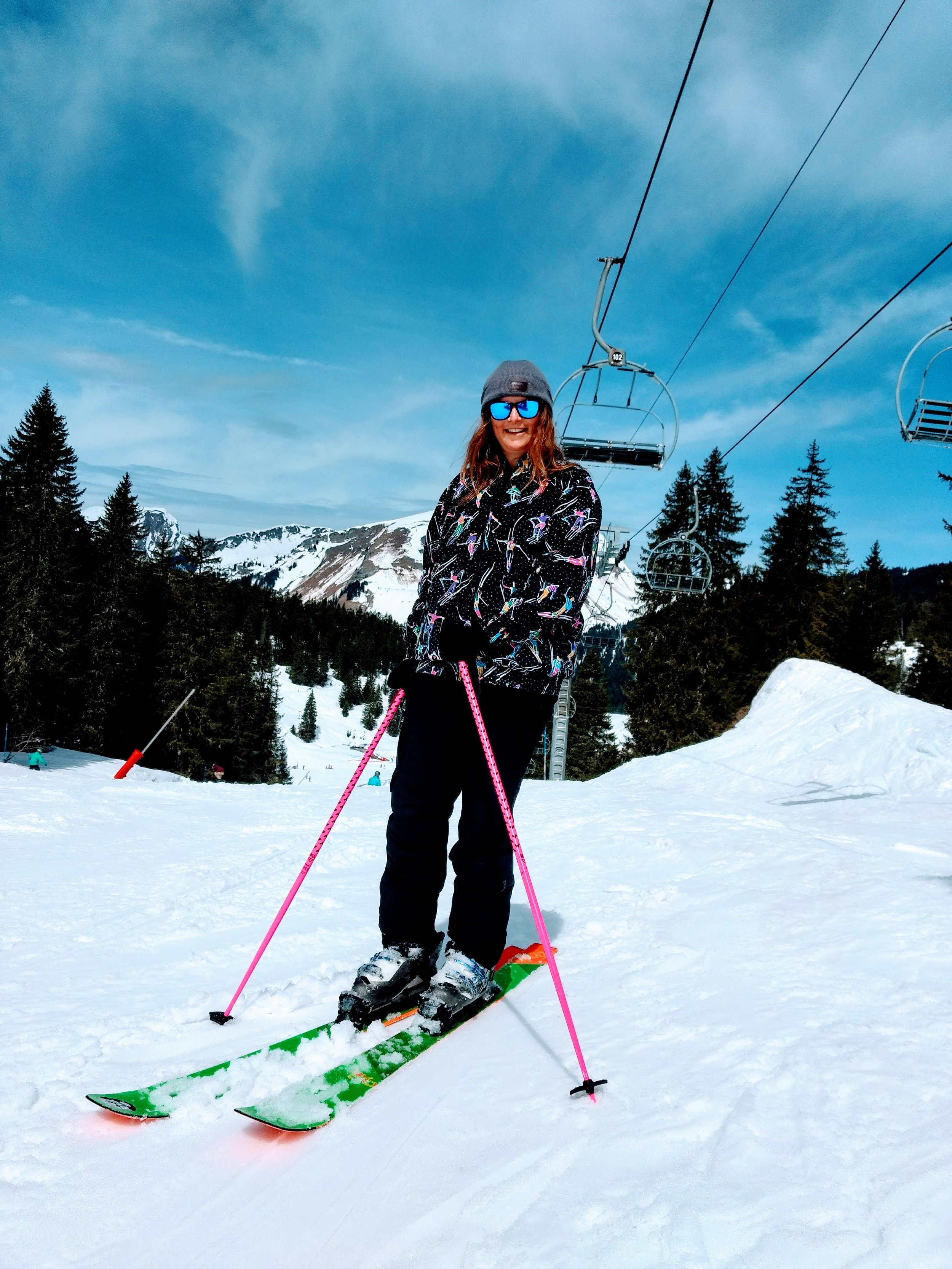 A woman wearing sunglasses, a gray beanie, a black jacket with colorful birds, black pants, and gloves, standing on skis on a snowy mountain slope, smiling. There are trees, ski lifts, and snow-covered mountains in the background.