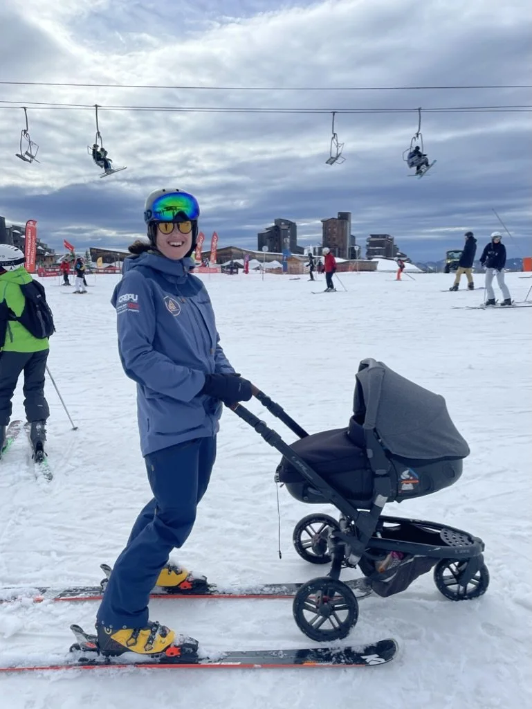 Woman in ski gear standing on skis and pushing a baby stroller on snow at a ski resort with ski lifts and other skiers in the background.