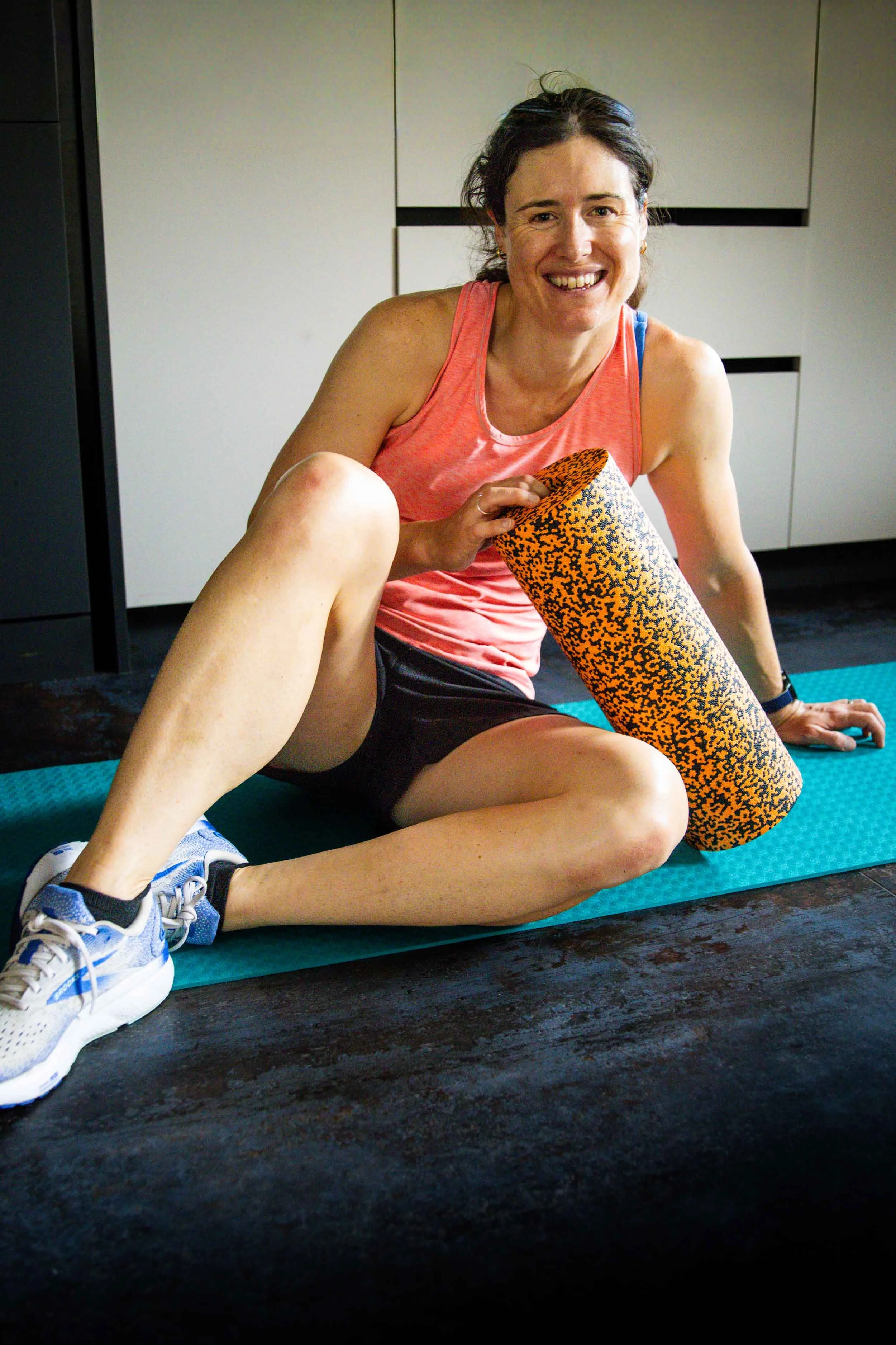 Woman in athletic clothes sitting on a yoga mat in a home gym, smiling while stretching her leg with a foam roller. She has dark hair tied back, and the room has white cabinets in the background.