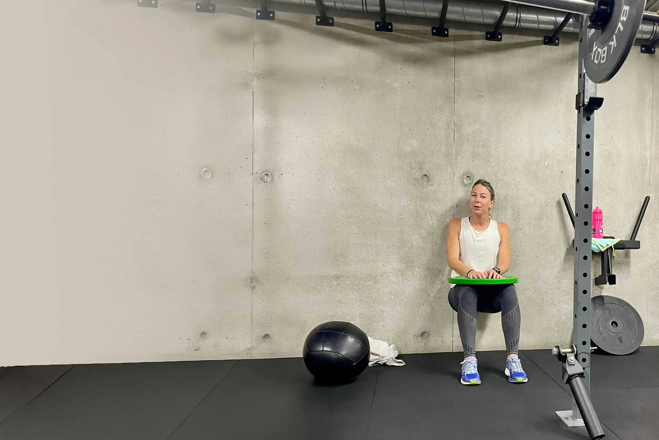 A woman sitting on a green exercise ball in a gym, surrounded by workout equipment including a pull-up bar, weight plates, and a pink water bottle. The woman is wearing workout clothes and smiling.