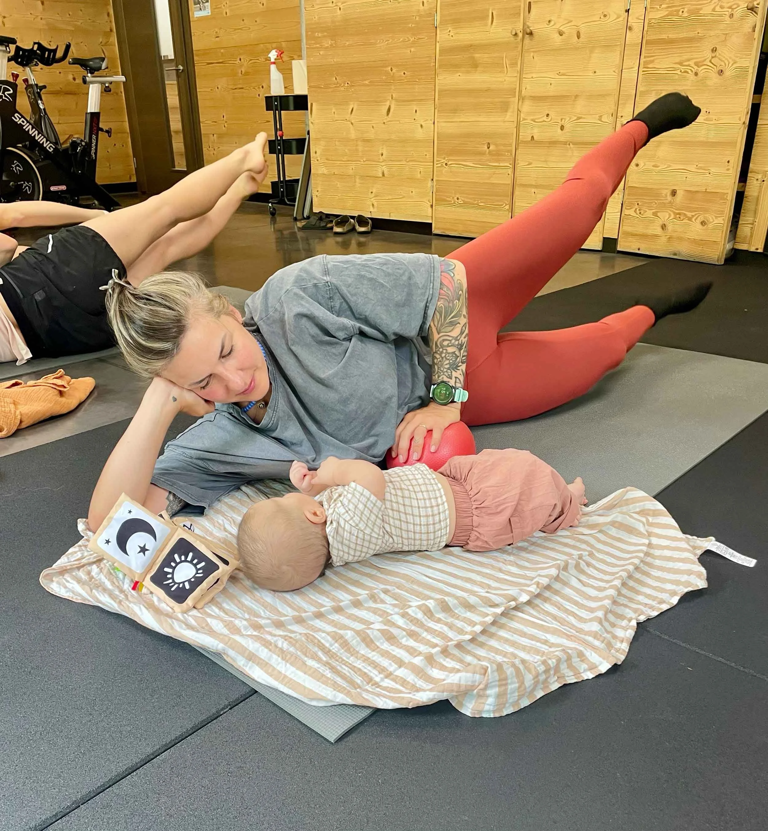 A woman in grey leggings lying on a striped mat, resting her head on her hand, with a baby lying next to her, and a woman in red leggings doing leg lifts nearby in a wooden-room exercise area.