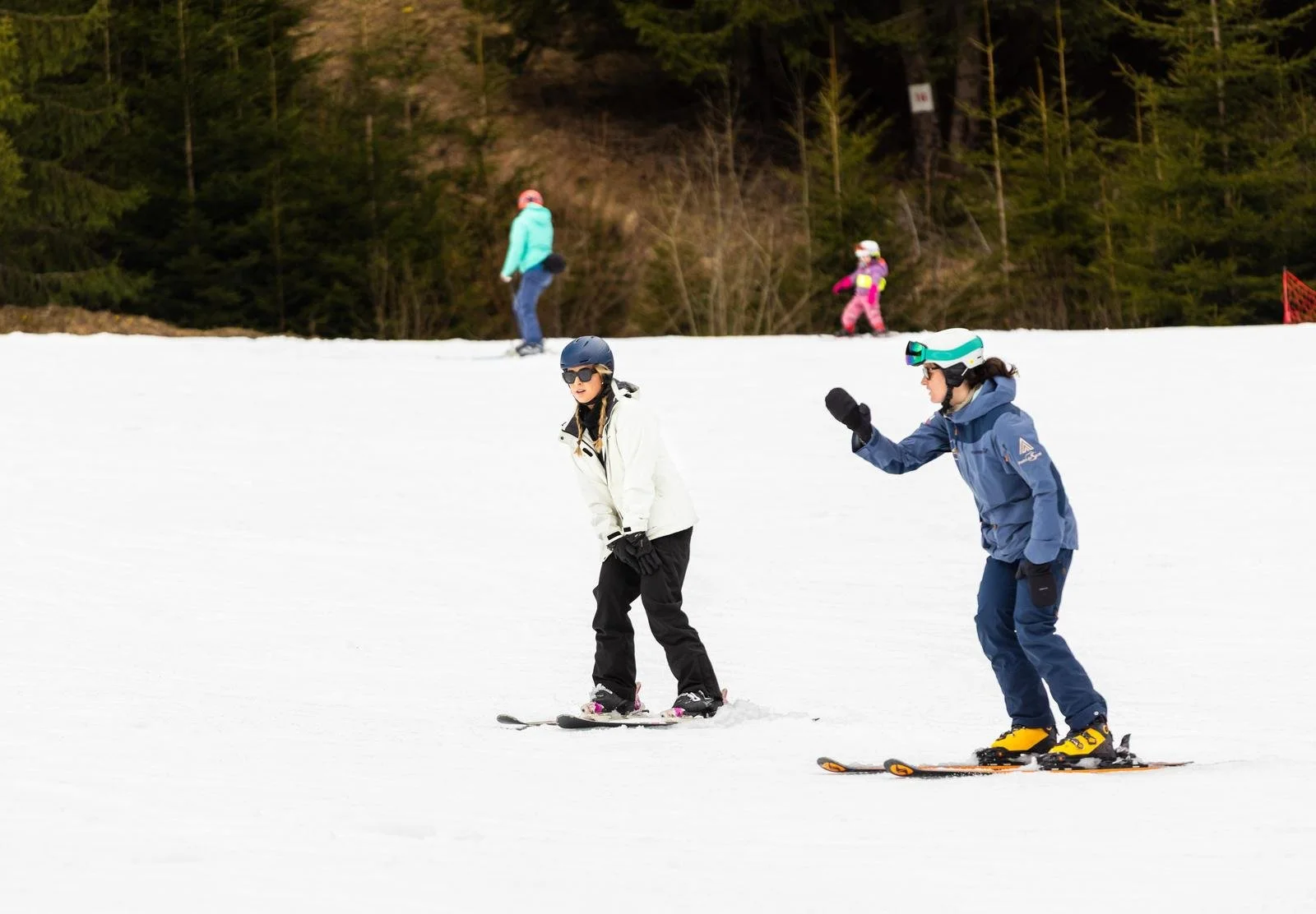 Two women skiing on snow, one wearing a white jacket and black pants, the other in a blue jacket and blue pants, both with helmets and goggles. Two children are skiing in the background, and a forest of trees is visible behind them.