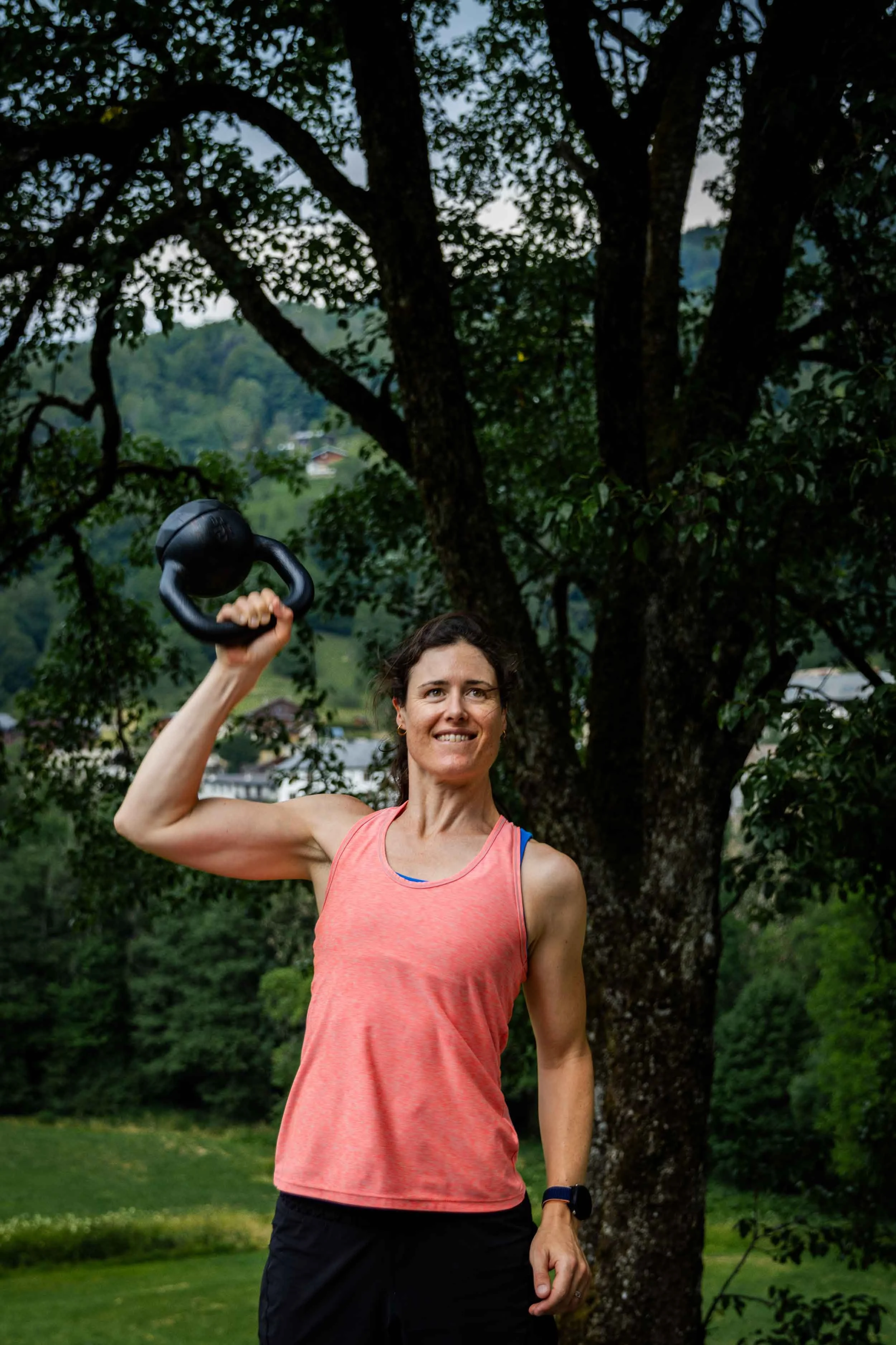 Woman in pink sleeveless workout top lifting a kettlebell outdoors in front of a large tree, with a scenic green landscape and houses in the background.