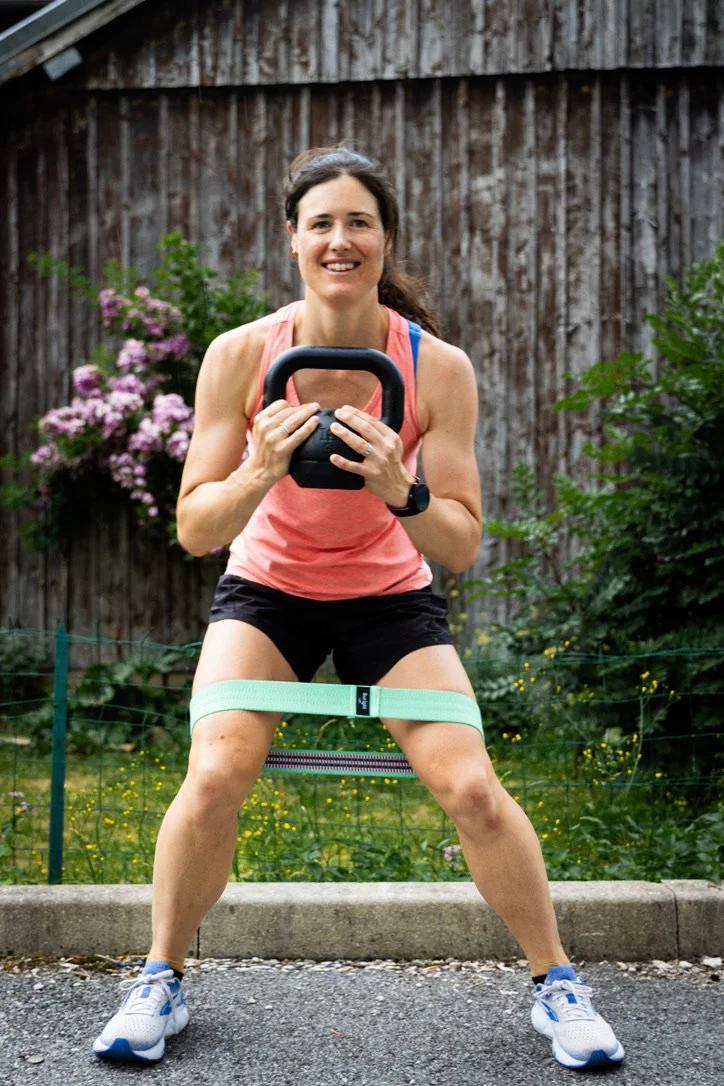 Woman exercising outdoors with a kettlebell, resistance band around her thighs, smiling, wearing athletic clothing, on a sidewalk with a wooden fence and greenery in the background.