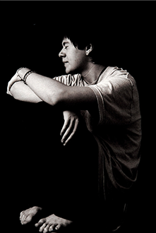A young person sitting and looking to the side in a thoughtful pose, wearing a casual shirt and bracelet, in black and white.