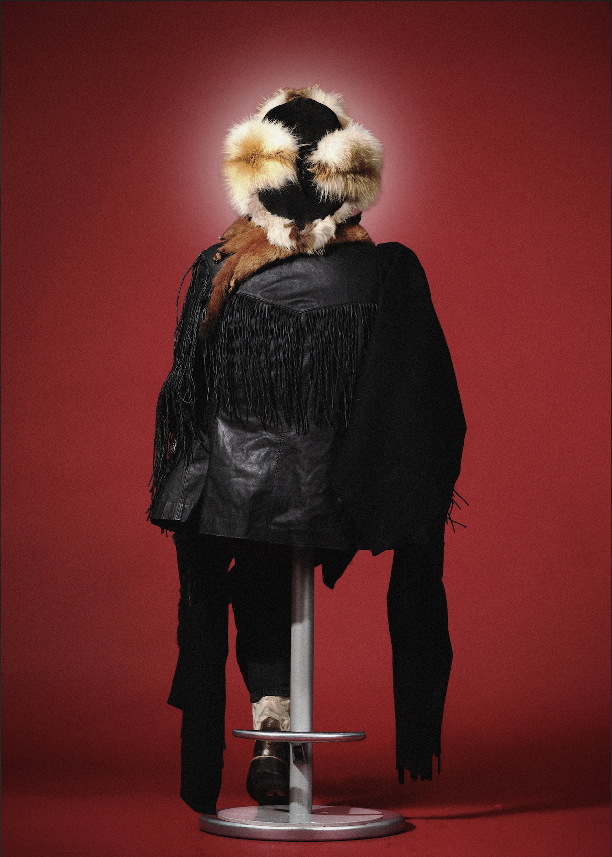 Person wearing a fur hooded coat with fringe details, standing behind a stool against a red background.