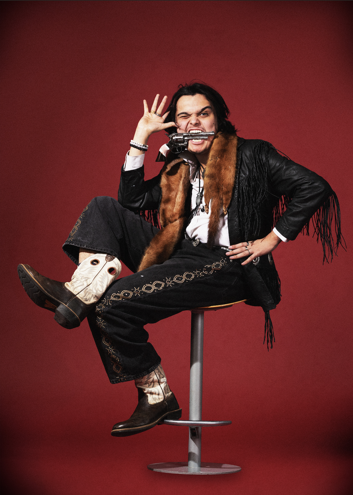 A person sitting on a stool against a red background, holding a silver revolver to their mouth, making a grimace, and wearing western-style clothing with cowboy boots, embroidered pants, a leather jacket with fringe, and a fur scarf.