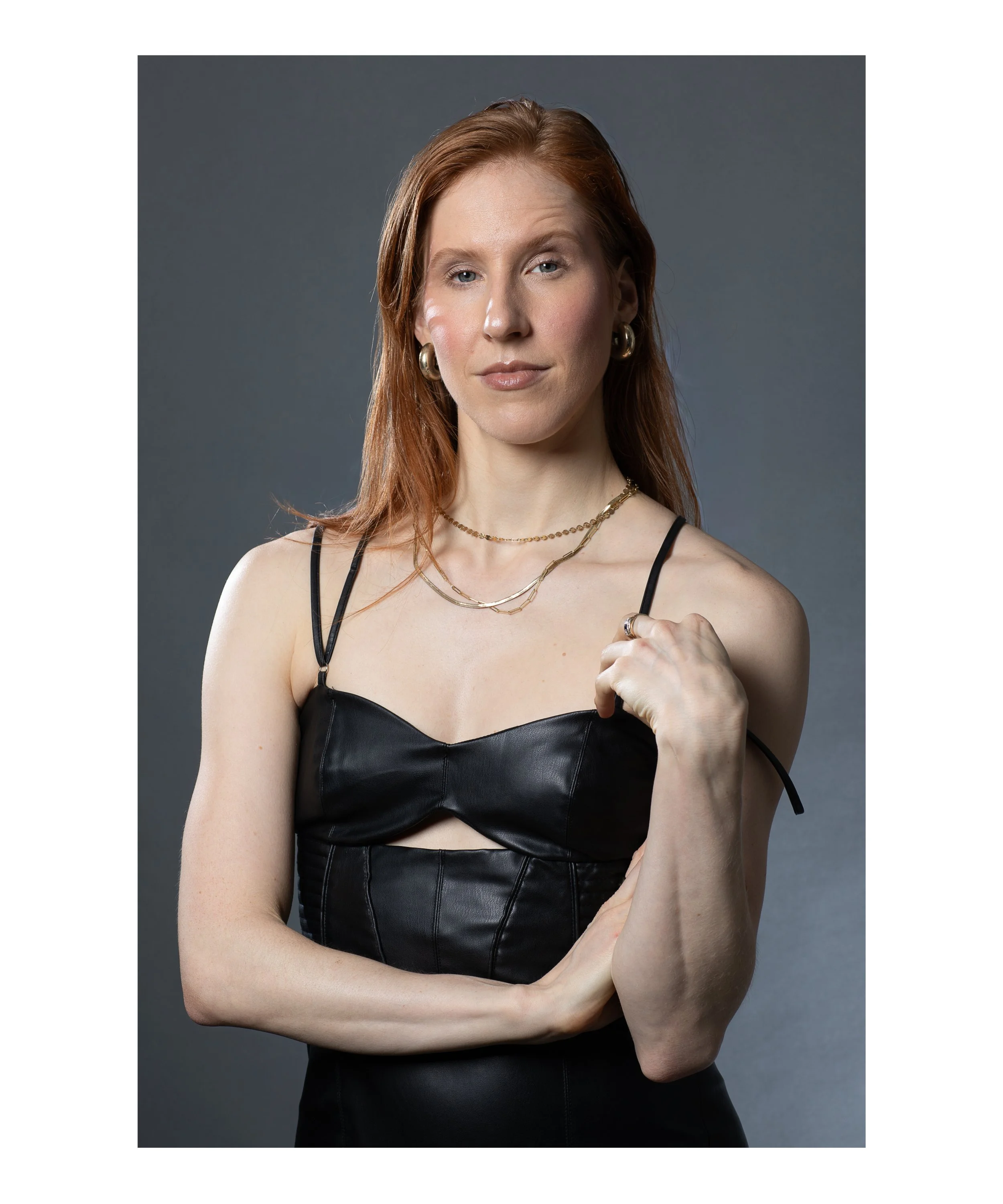 A woman with red hair wearing a black leather top, layered gold necklaces, hoop earrings, and a ring on her finger, posing against a dark gray background.
