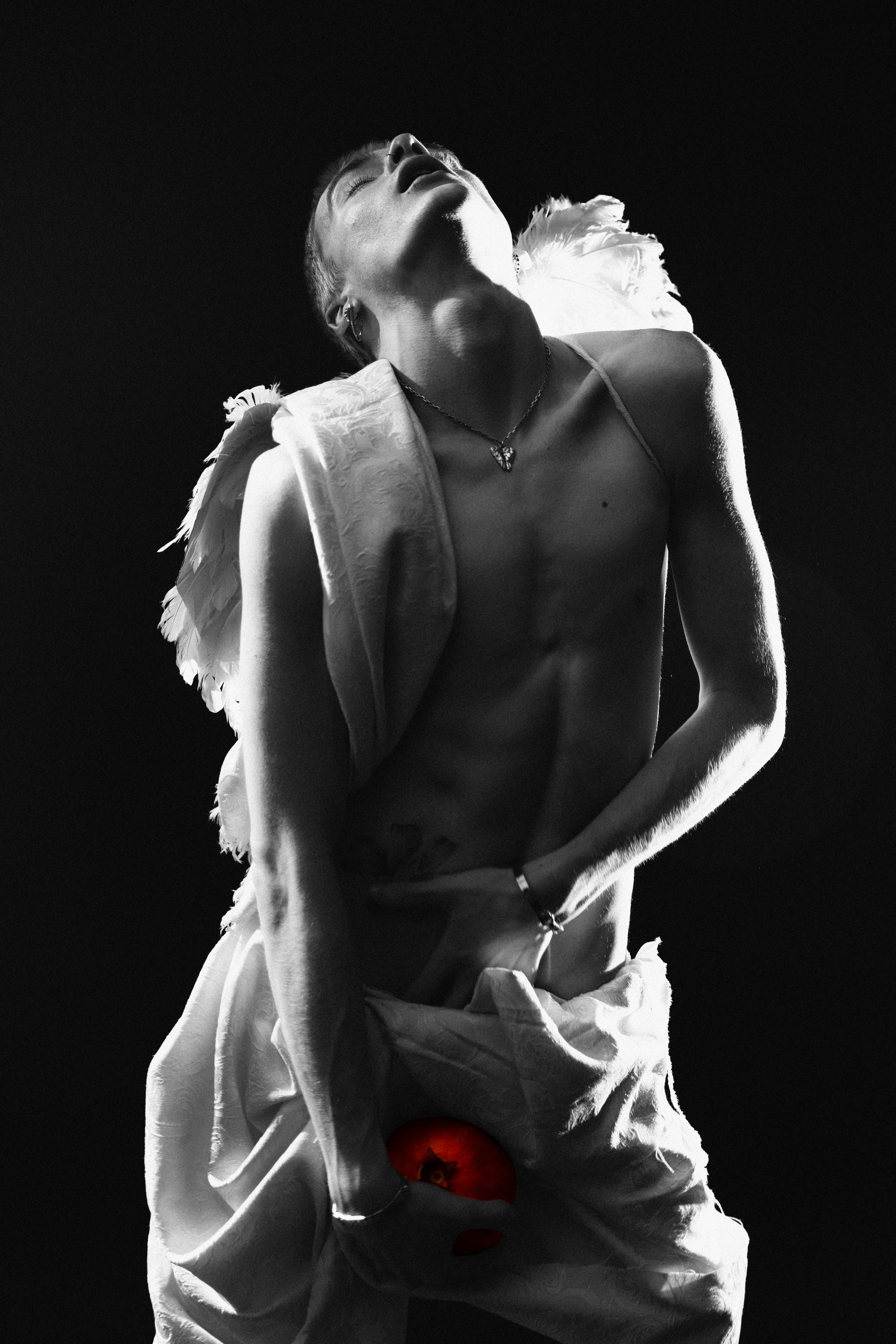 A black and white photo of a person with short hair, closed eyes, and an open mouth, wearing earrings, a necklace, and a feathered shawl. They are topless, with a cloth wrapped around their waist, and a red apple is photoshopped into their groin area.