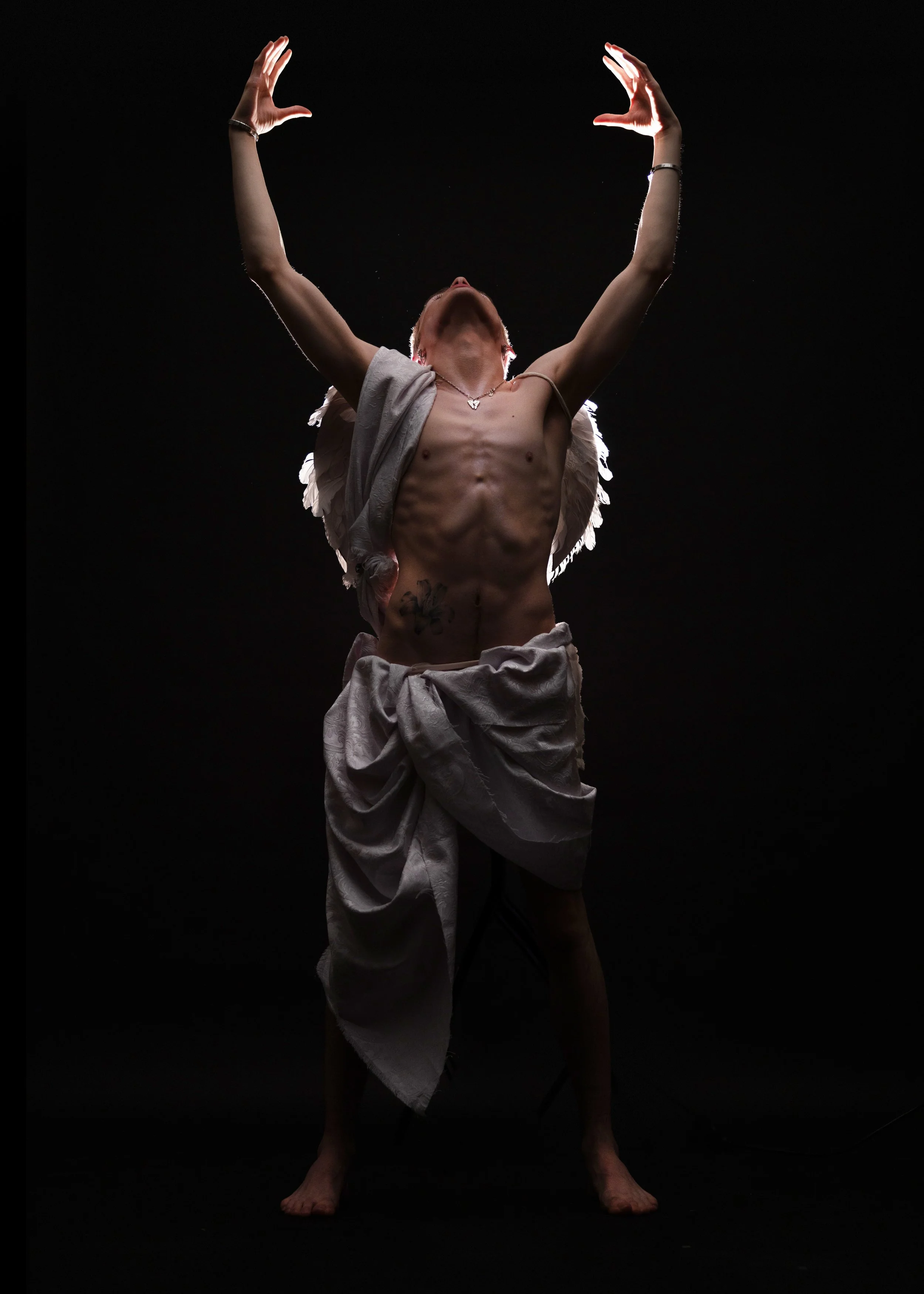 Person with angel wings, shirtless, wearing draped fabric around waist, standing with arms raised, head tilted back, illuminated against a dark background.