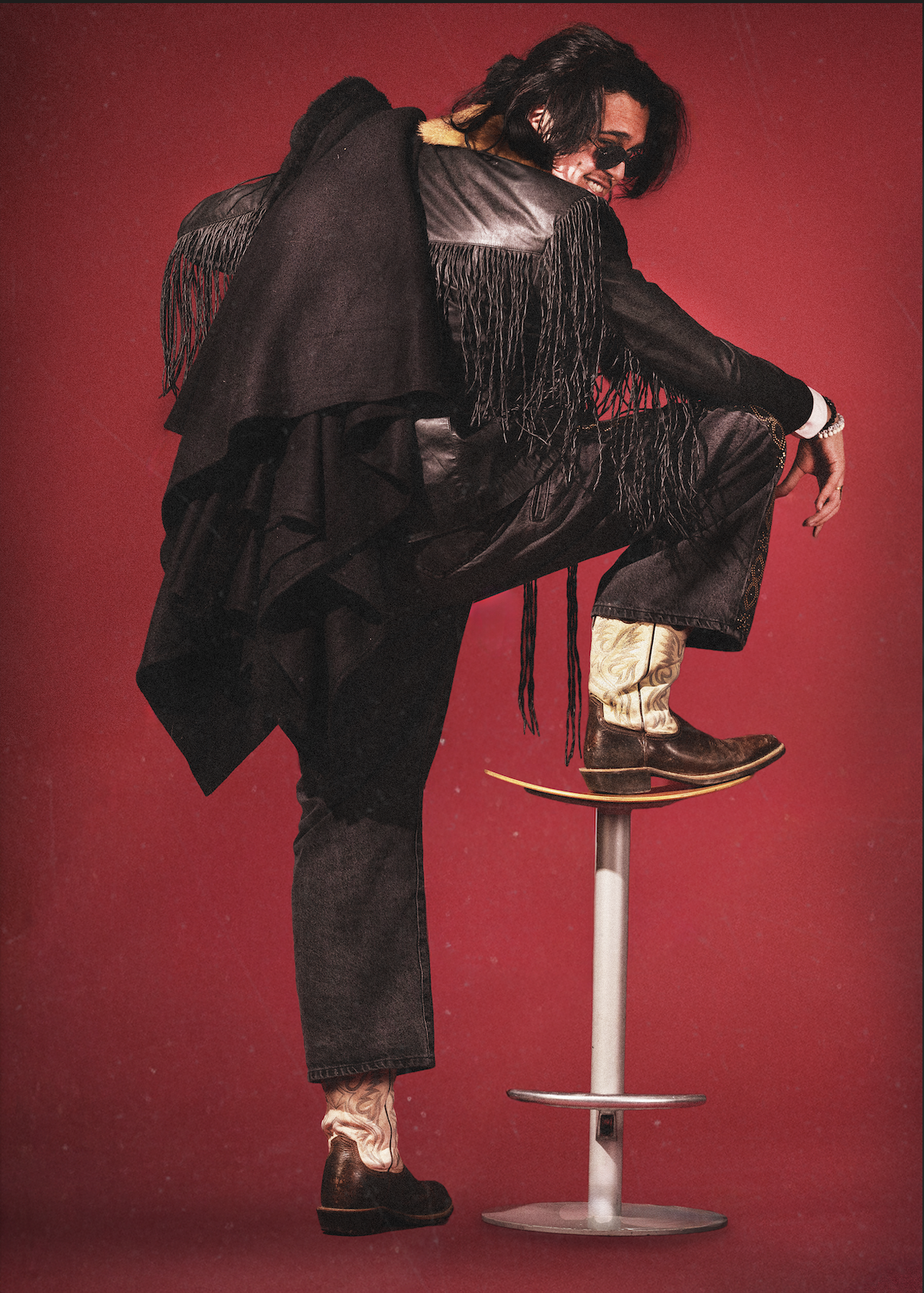 A person with long dark hair, wearing sunglasses, a black leather jacket with tassels, black pants, and cowboy boots, is striking a pose with one foot raised on a stool against a red background.