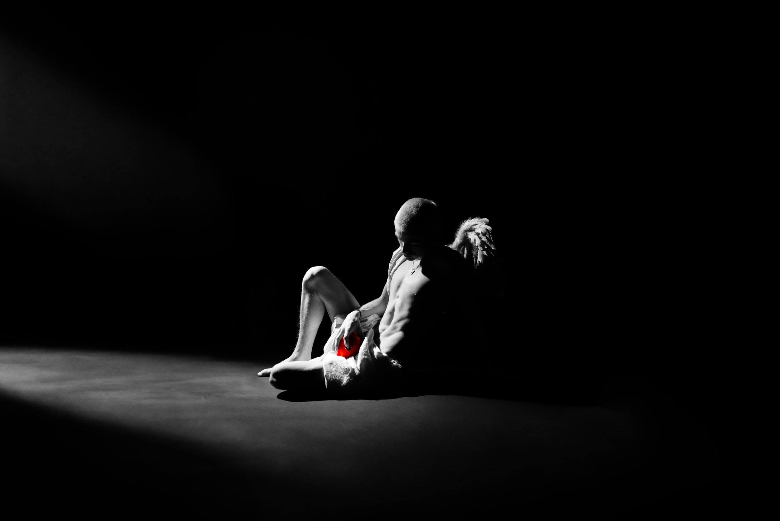 A black and white photo of a young person sitting on the floor, illuminated by a spotlight, with a white dog lying next to them, holding a red plush toy.