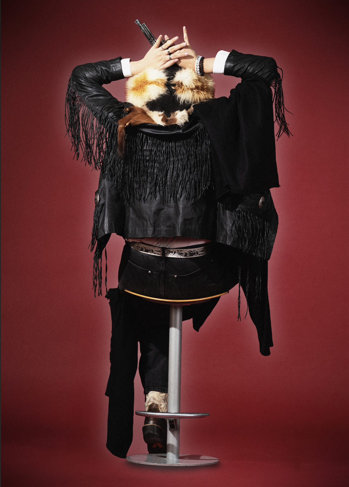 A person with a painted face, wearing a furry hat with a fur collar, a black fringed leather jacket, and black jeans, sitting on a stool against a red background, with their hands behind their head and their face partially obscured.