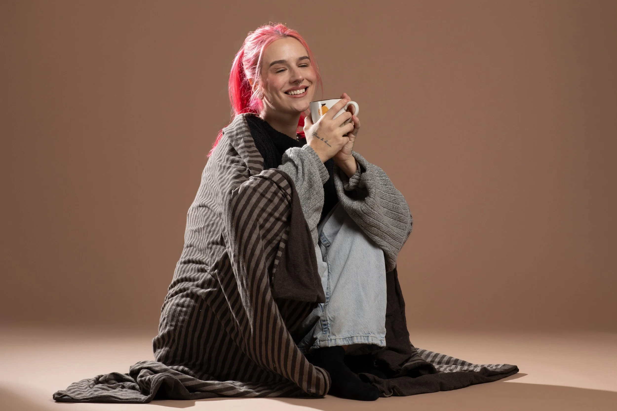 A young woman with pink hair smiling and holding a cup, sitting on the floor with her knees up, against a plain beige background.