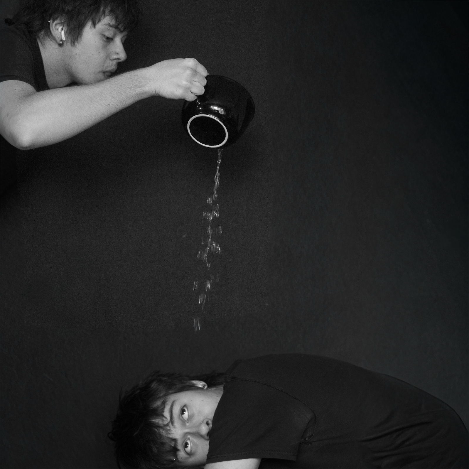 A person with short dark hair and multiple ear piercings leans over a black surface, with water pouring from a black cup onto their back. The person looks directly at the camera.