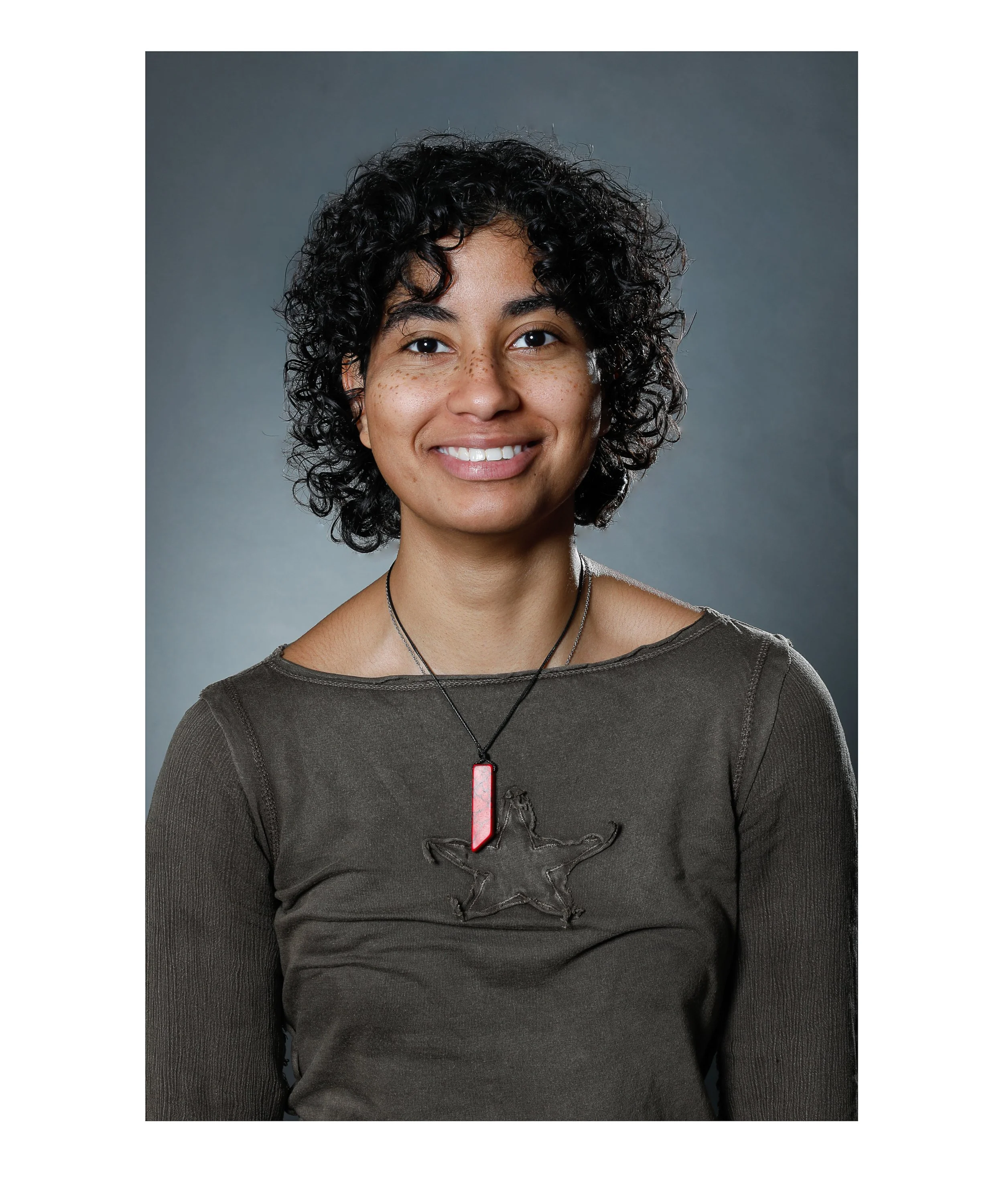 A woman with short, curly dark hair, light brown skin, and freckles on her face, smiling at the camera. She wears a dark gray long-sleeve shirt with a graphic of a ram's head and a necklace with a rectangular red pendant.