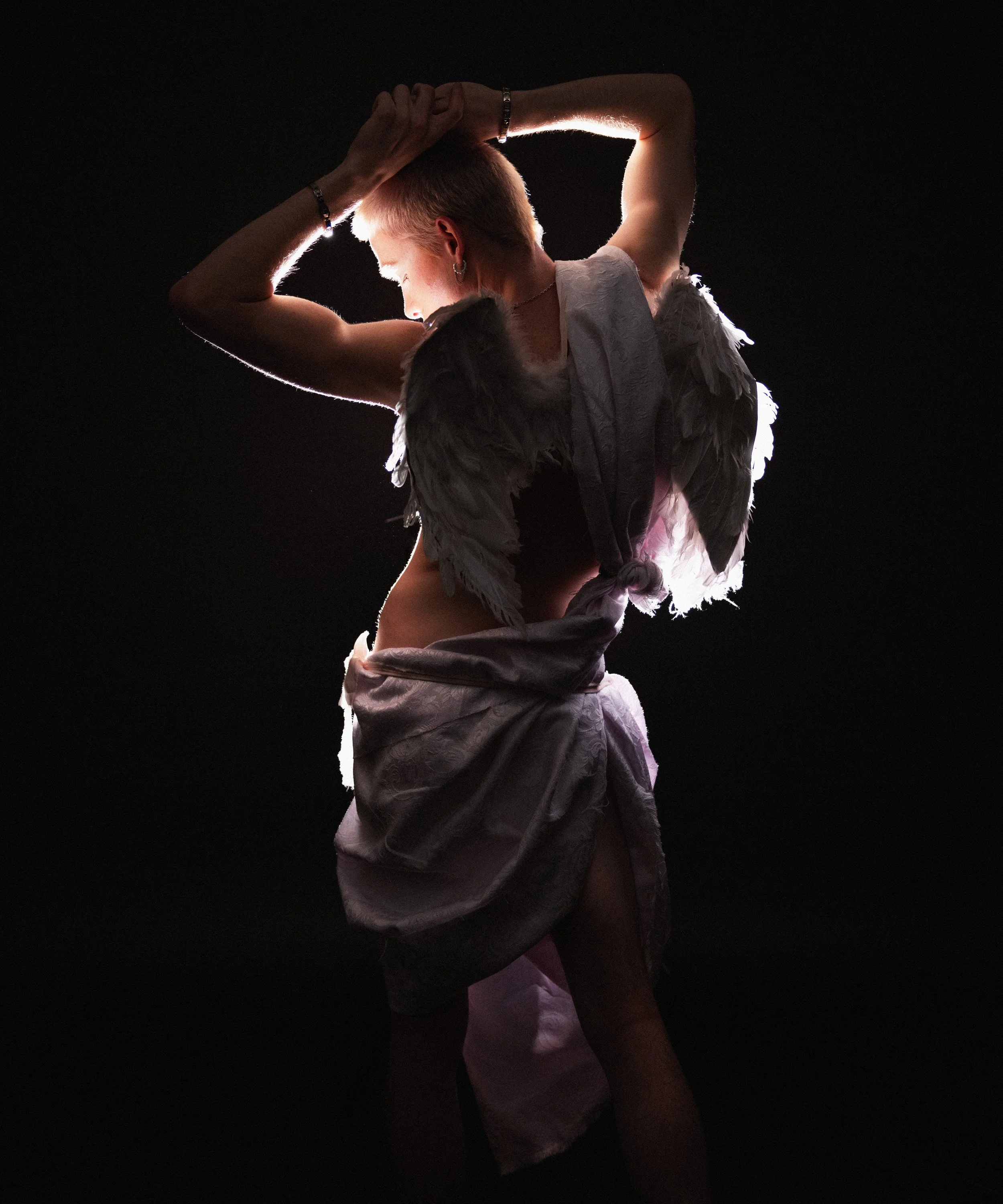 A person with short blonde hair and earrings, wearing a white shirt with feathered sleeves, is posed with hands behind their head against a black background, illuminated from the back.