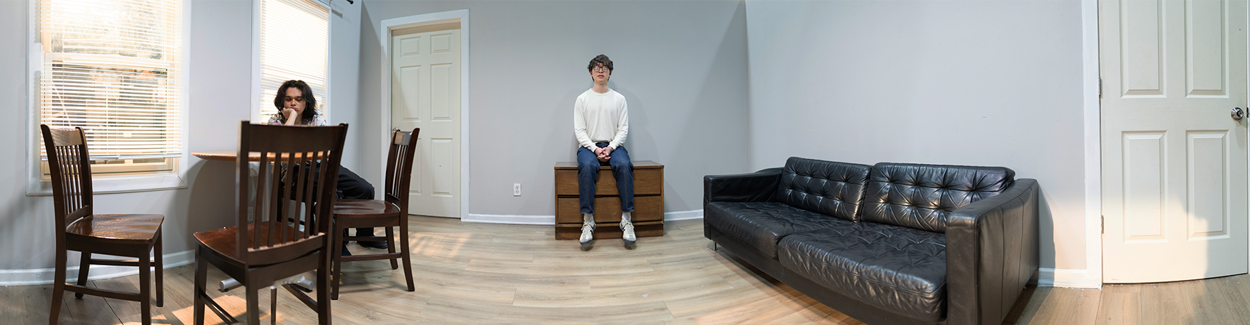 A living room with a wooden dining table and four chairs on the left, a woman sitting at the table with her hand on her face, a person standing on a piece of furniture in the center, and a black leather couch on the right. The room has light-colored walls, a wooden floor, and two windows with blinds.