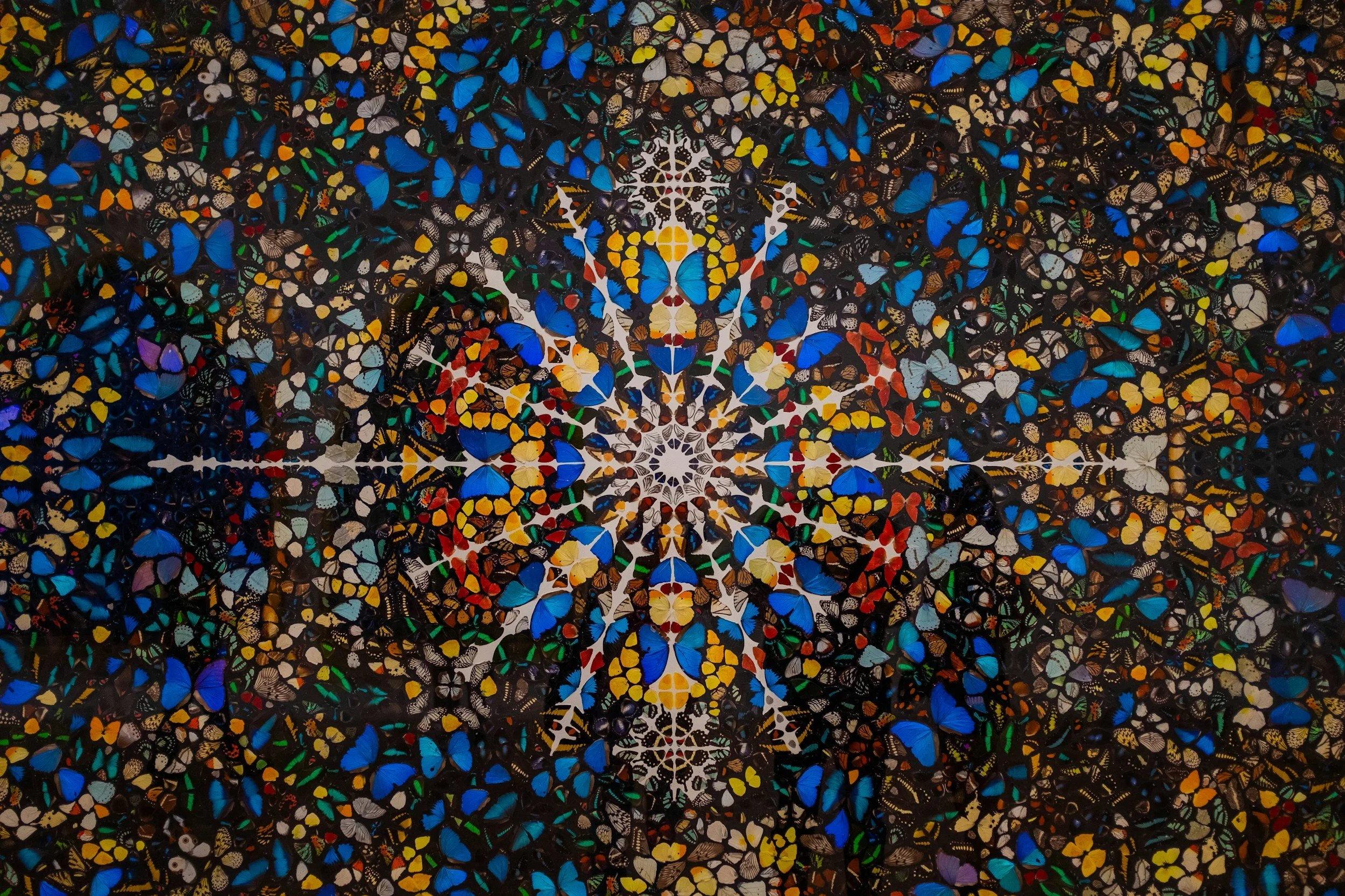 A large mosaic artwork composed of numerous butterflies, predominantly in blue, yellow, orange, white, and black, arranged in a symmetrical pattern with intricate details.