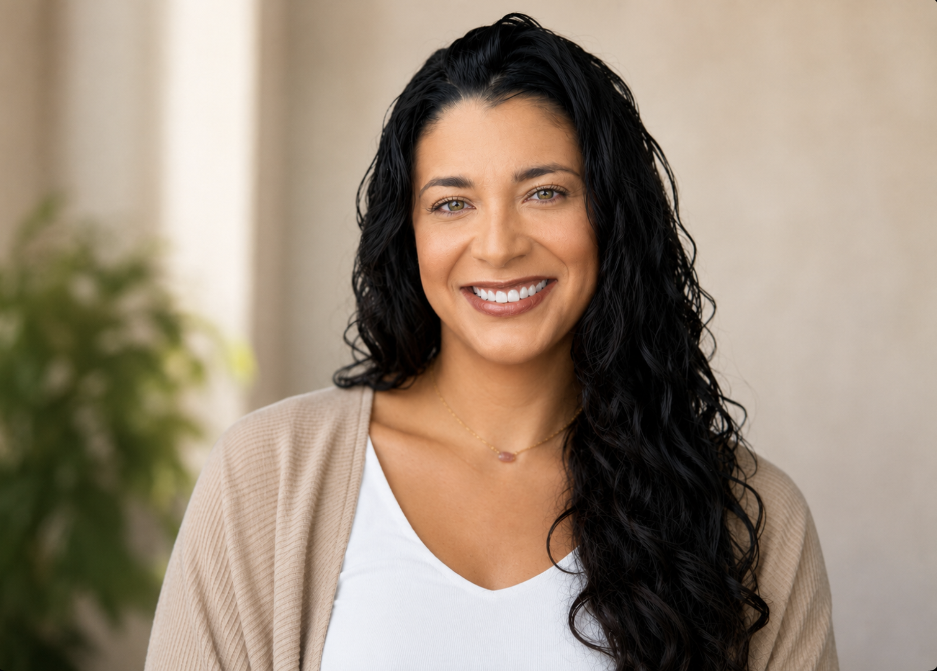 A smiling woman with long black curly hair, wearing a beige cardigan over a white top, and a gold necklace with a small pendant, in a well-lit indoor setting.