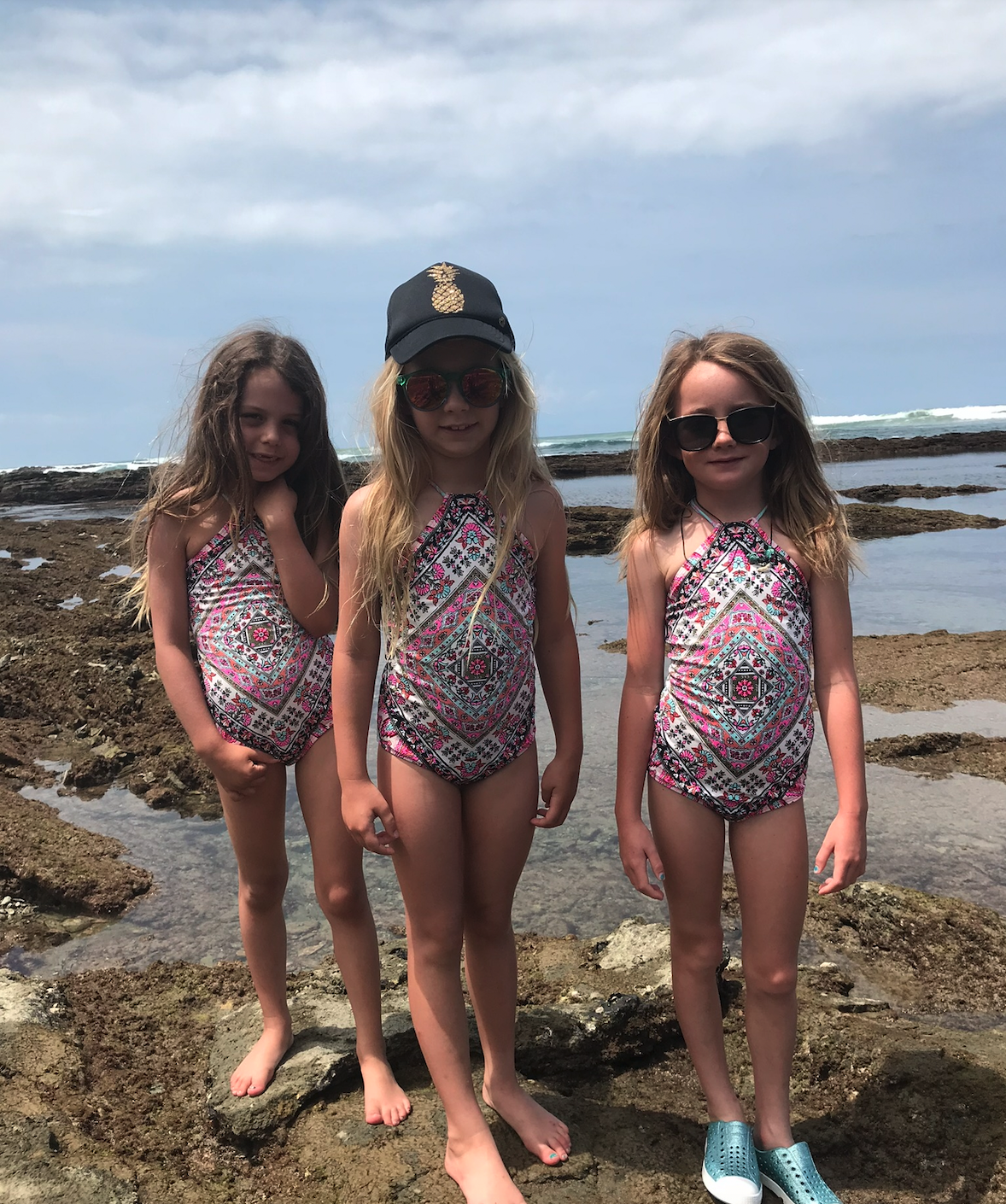 Three young girls standing on rocks near the ocean, wearing swimsuits with geometric patterns, sunglasses, and one wearing a hat.