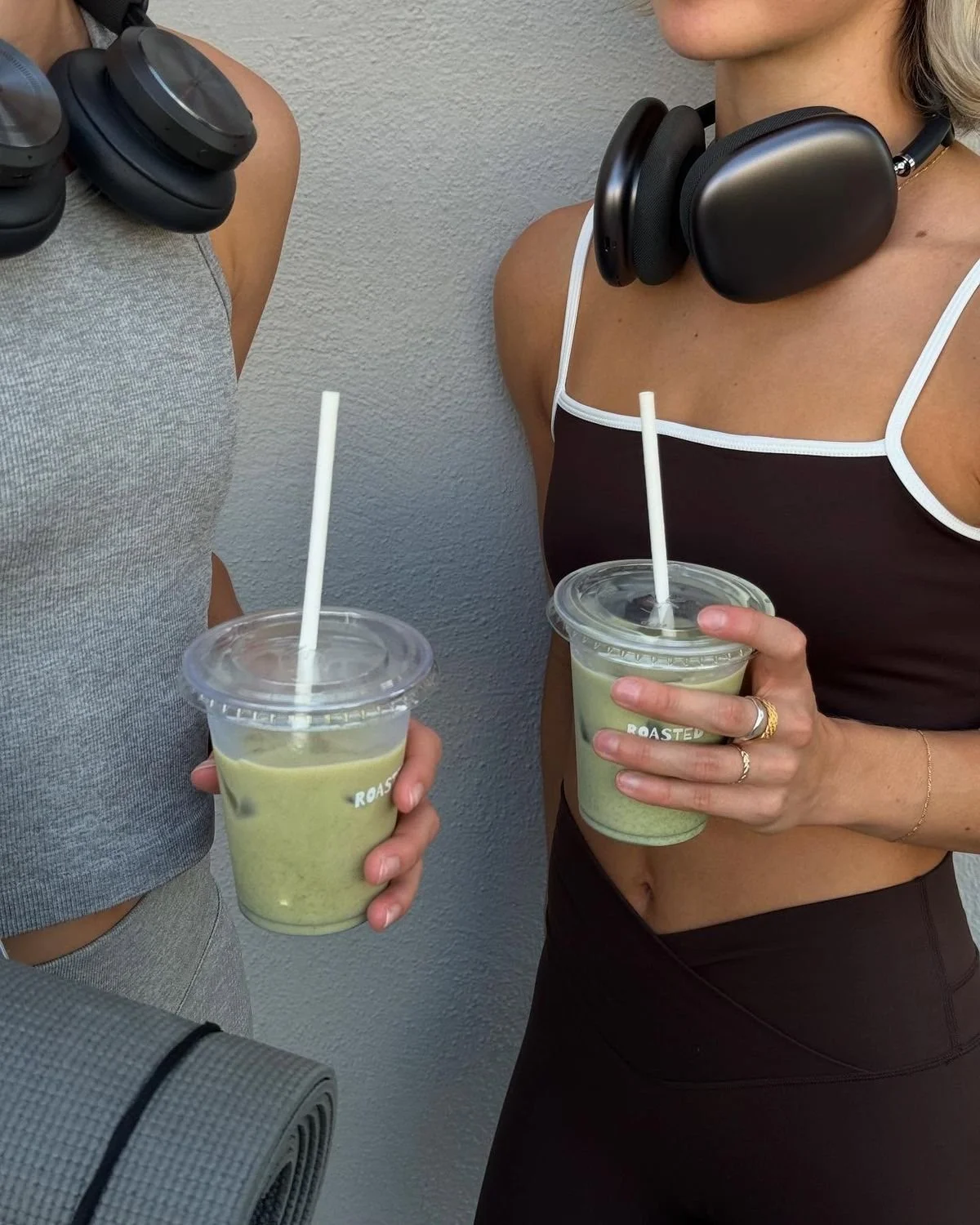 Two women outdoors holding cups of green smoothie with straws, wearing athletic clothing and headphones around their necks.