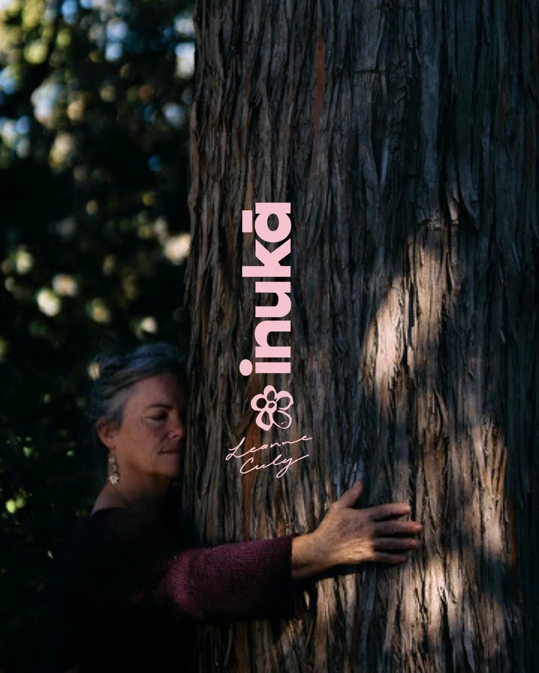 A woman with short gray hair, wearing earrings and a maroon sweater, embraces a large tree trunk in a forest. Sunlight filters through the dark green foliage, creating shadows and a peaceful atmosphere.
