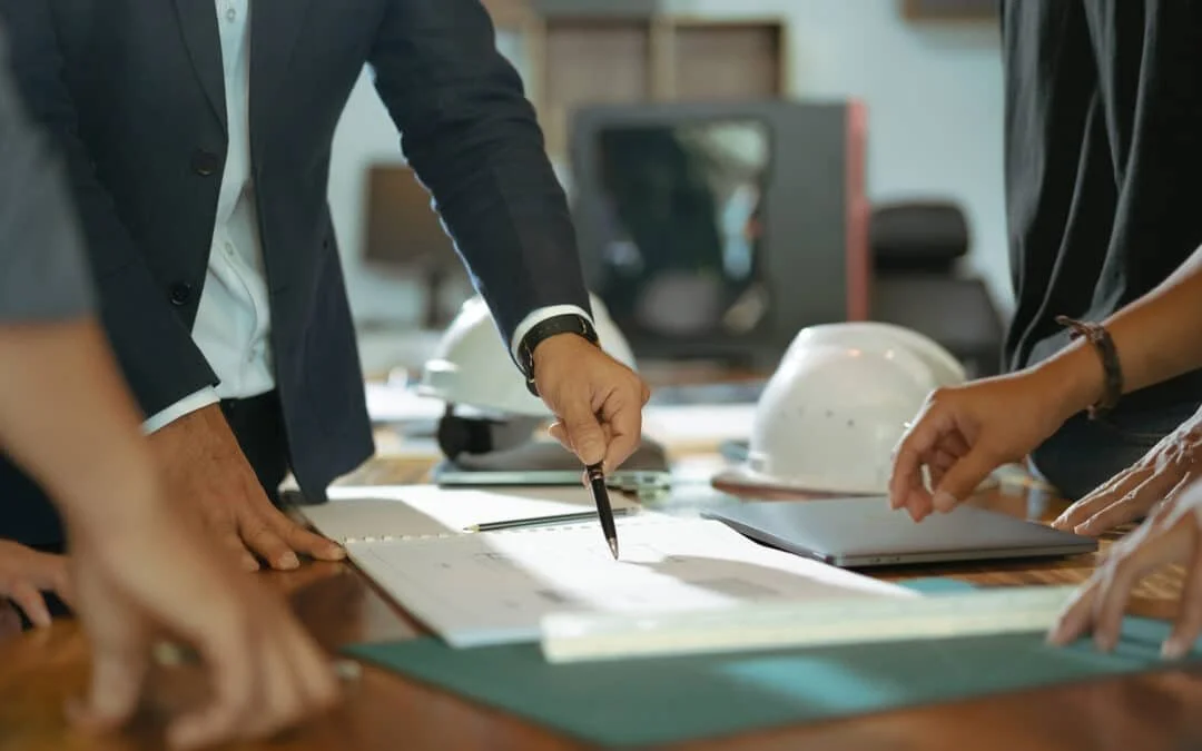 People examining blueprints or architectural plans on a table in a workspace, with safety helmets and laptops nearby.