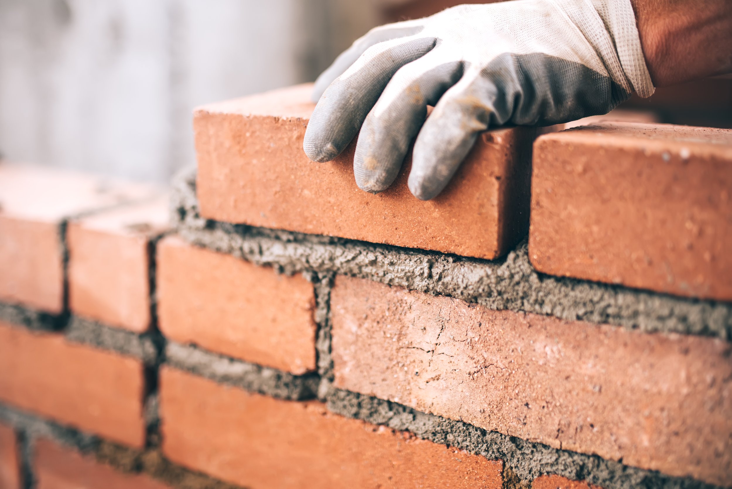 Close-up of a worker's hand wearing a gray glove, laying bricks in a row during bricklaying construction.