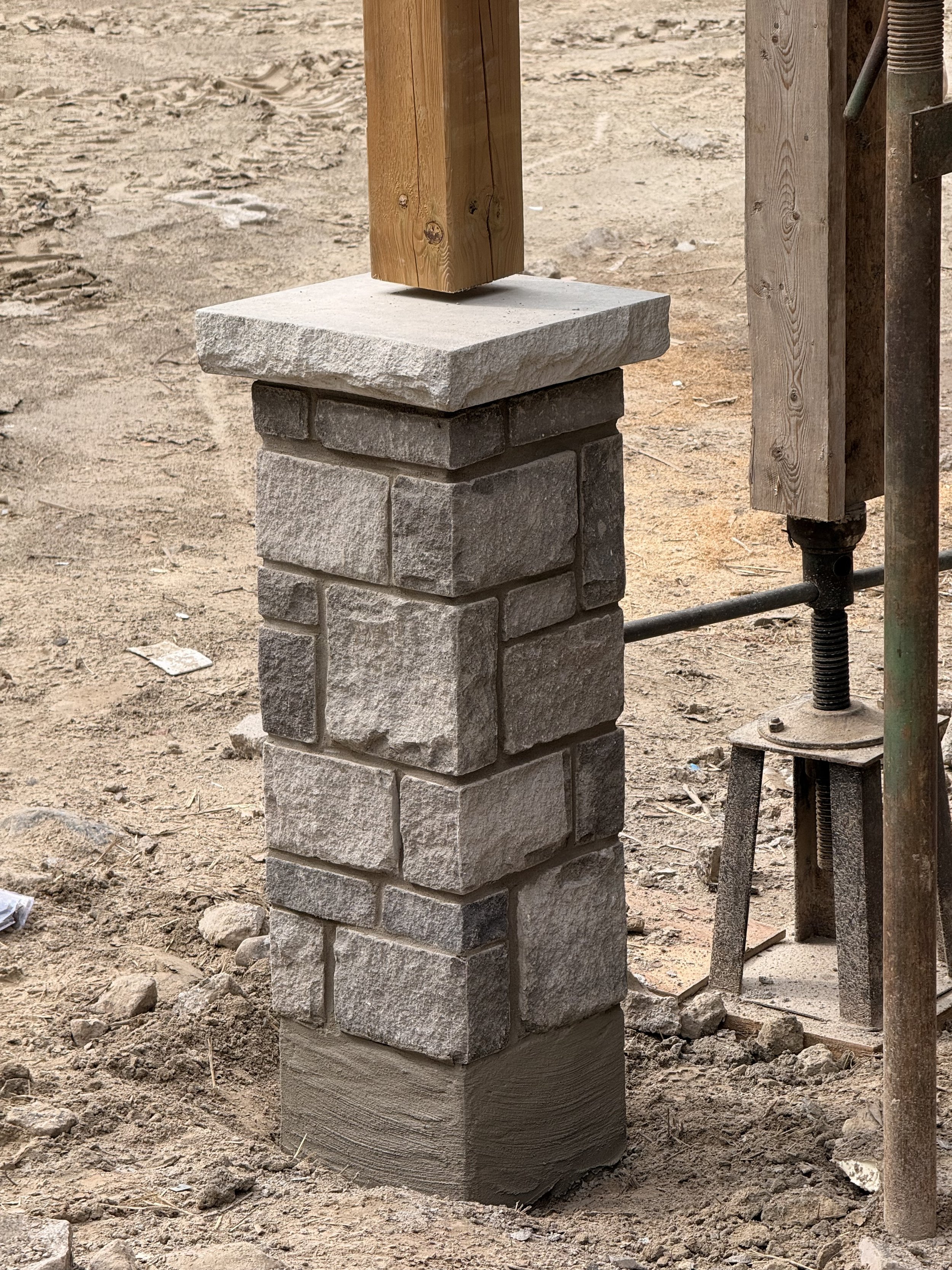 A stone pillar with a wooden beam on top, under construction, with dirt ground and construction equipment nearby.