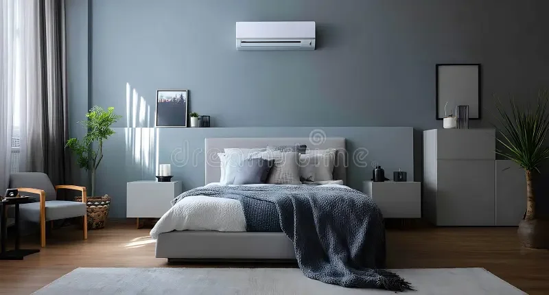 Modern bedroom with gray walls, a large bed with gray and white bedding, an air conditioner above the bed, two nightstands with lamps, framed art, a gray dresser with decor, a plant in a basket, and a gray armchair by the window.
