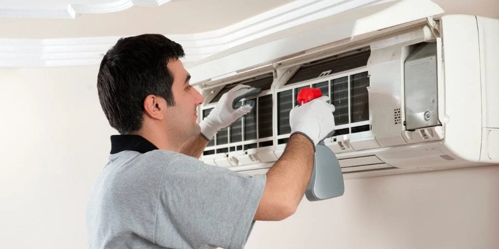 A man cleaning an air conditioner unit mounted on a wall with a spray bottle and cloth.