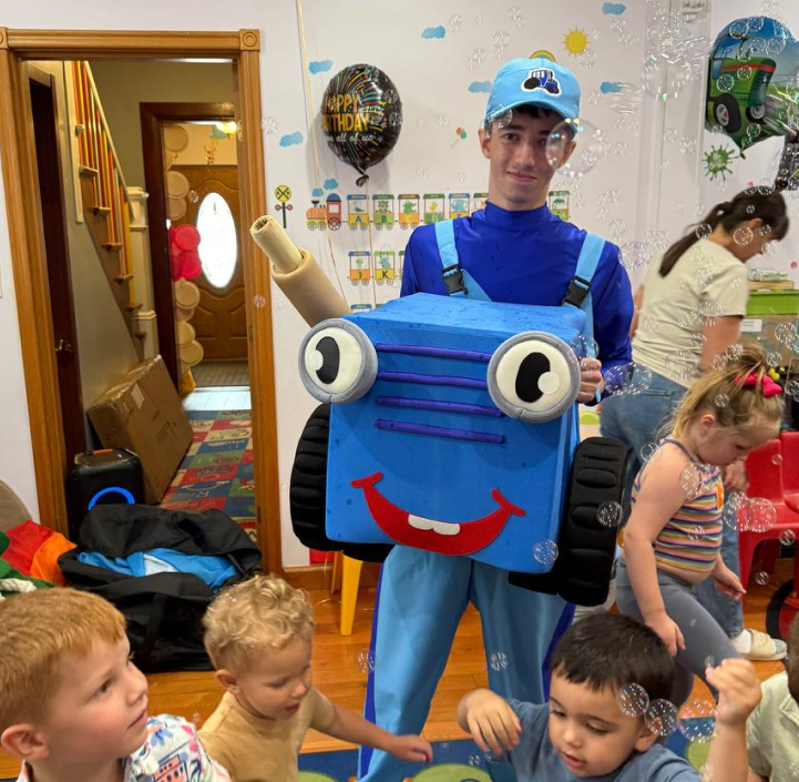 A young man dressed as a robot with a boxy blue costume, large googly eyes, and a red smile, standing among children playing at a birthday party indoors.
