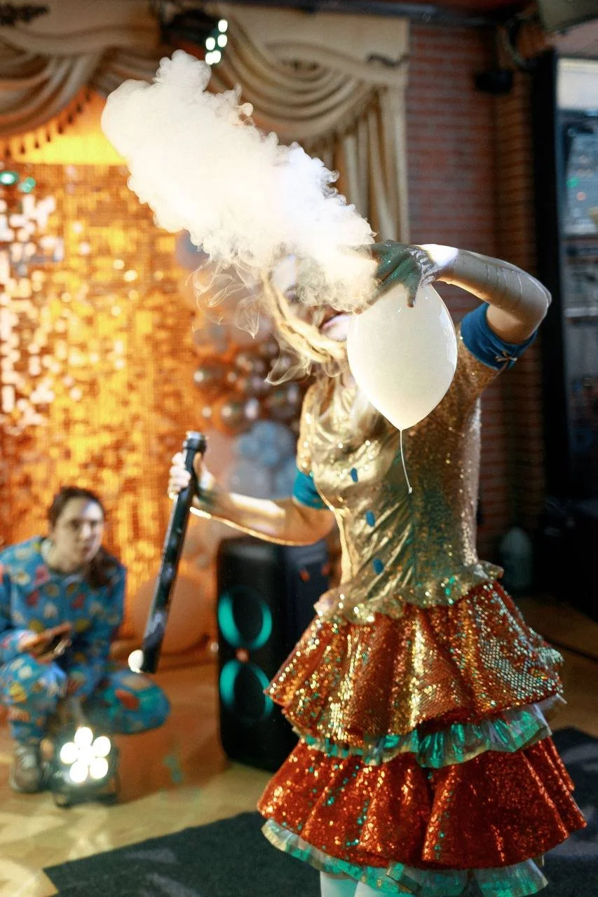 A performer dressed in a gold and red sequined costume holding a balloon and emitting smoke in a decorated indoor setting, with a person in colorful clothing squatting in the background.