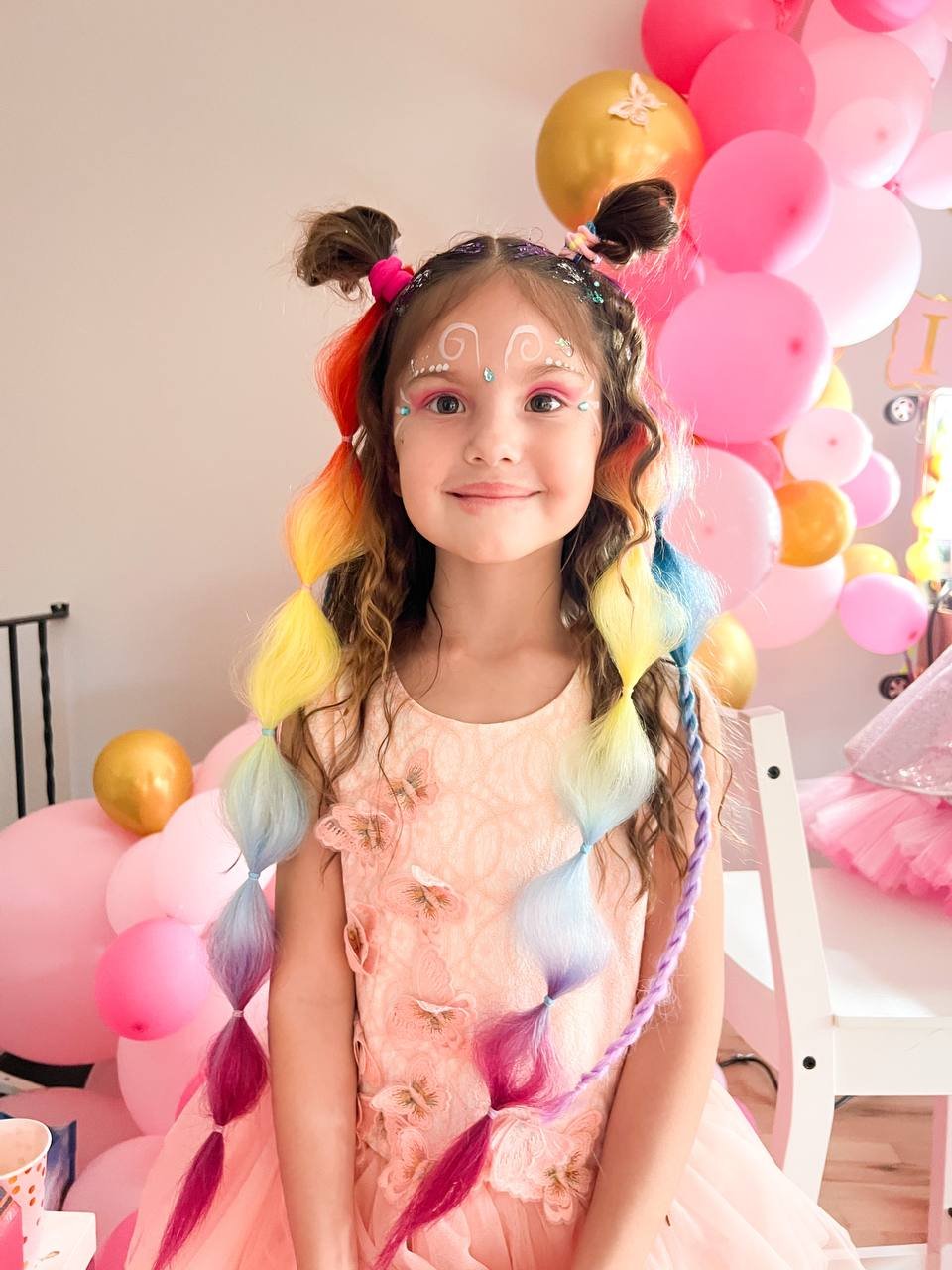 Child girl with rainbow-colored hair in pigtails, rainbow makeup, smiling at a celebration with pink and gold balloons in the background.
