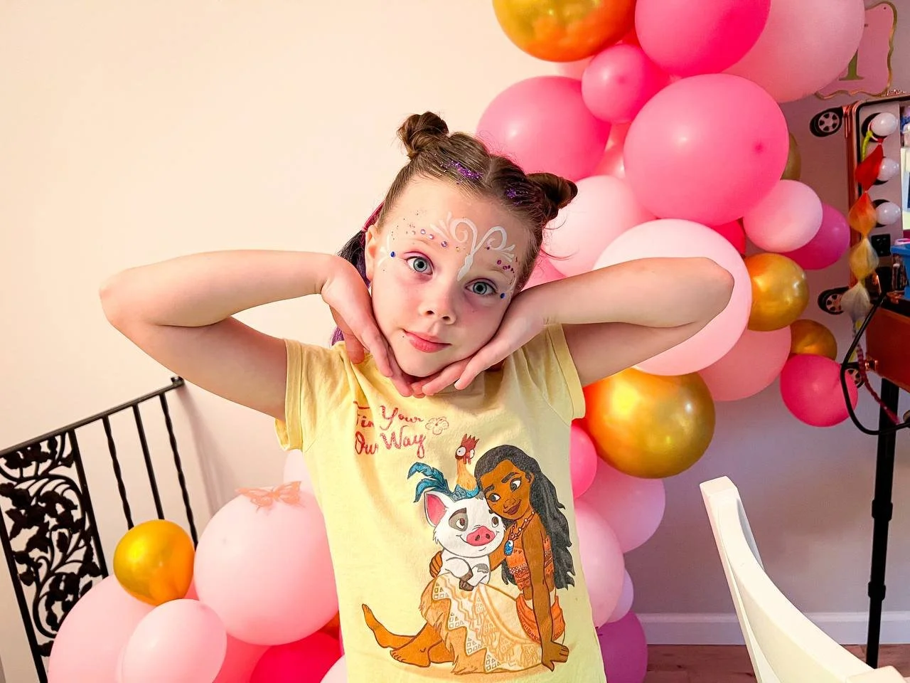 A young girl posing with pink, gold, and peach balloons in the background, wearing a yellow shirt featuring characters from Disney's Moana, with face makeup and her hair styled in two buns.