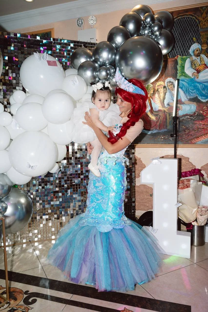 A woman dressed as a mermaid with a red wig and a party hat holds a young girl in a white dress at a birthday celebration with balloons and decorations.