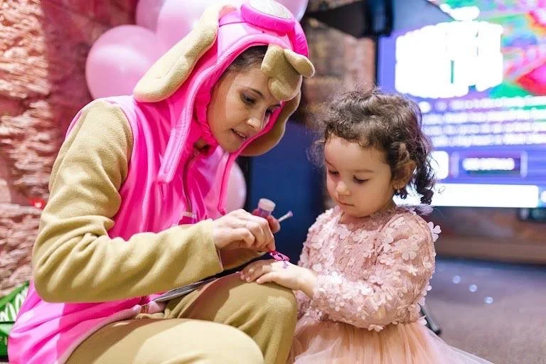 Adult dressed in a pink and beige animal costume giving a manicure to a young girl in a pink dress, in a festive setting with balloons and decorations.