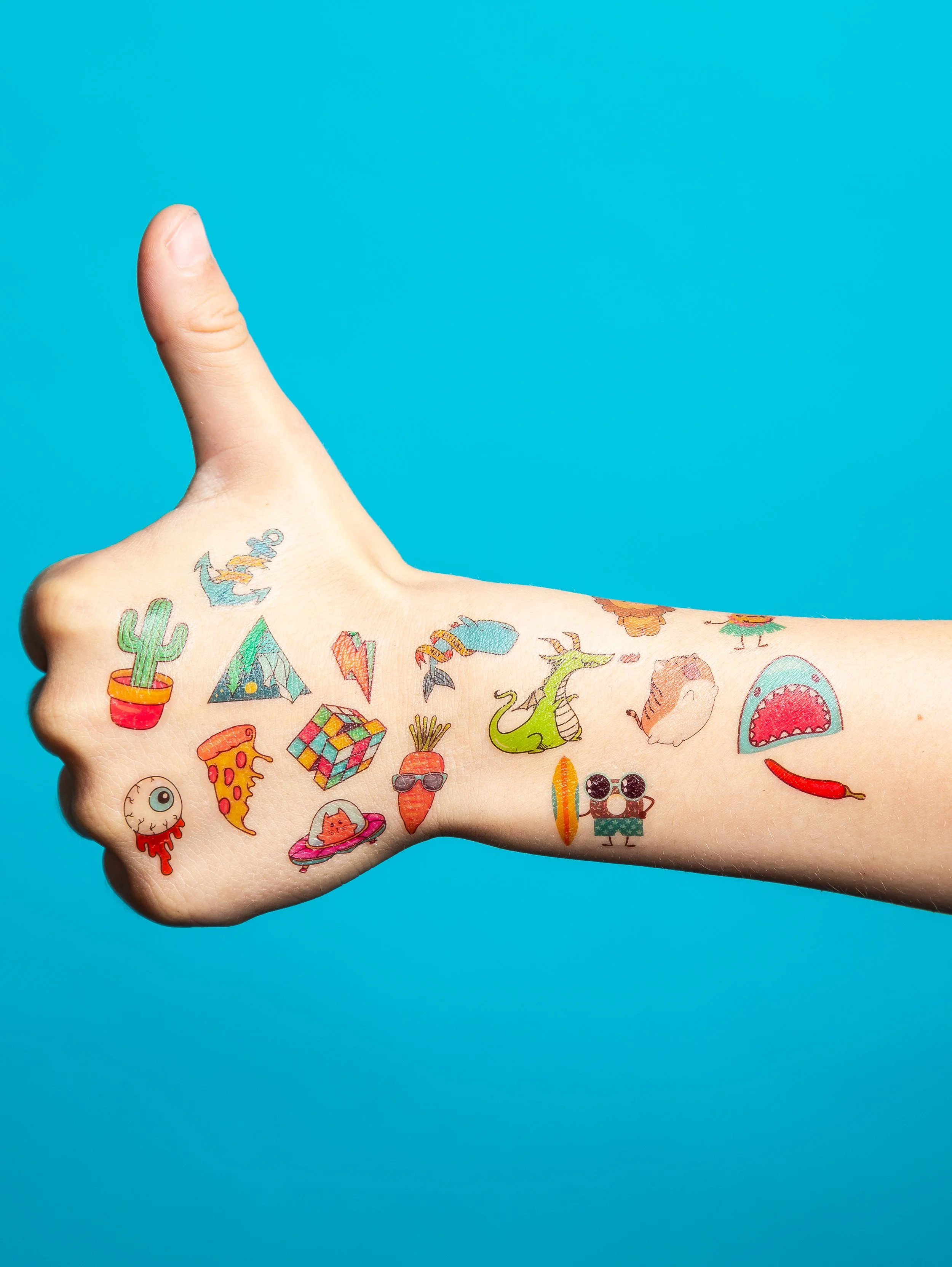 A person's arm with colorful tattoos of various objects and cartoon characters, giving a thumbs-up gesture against a solid blue background.