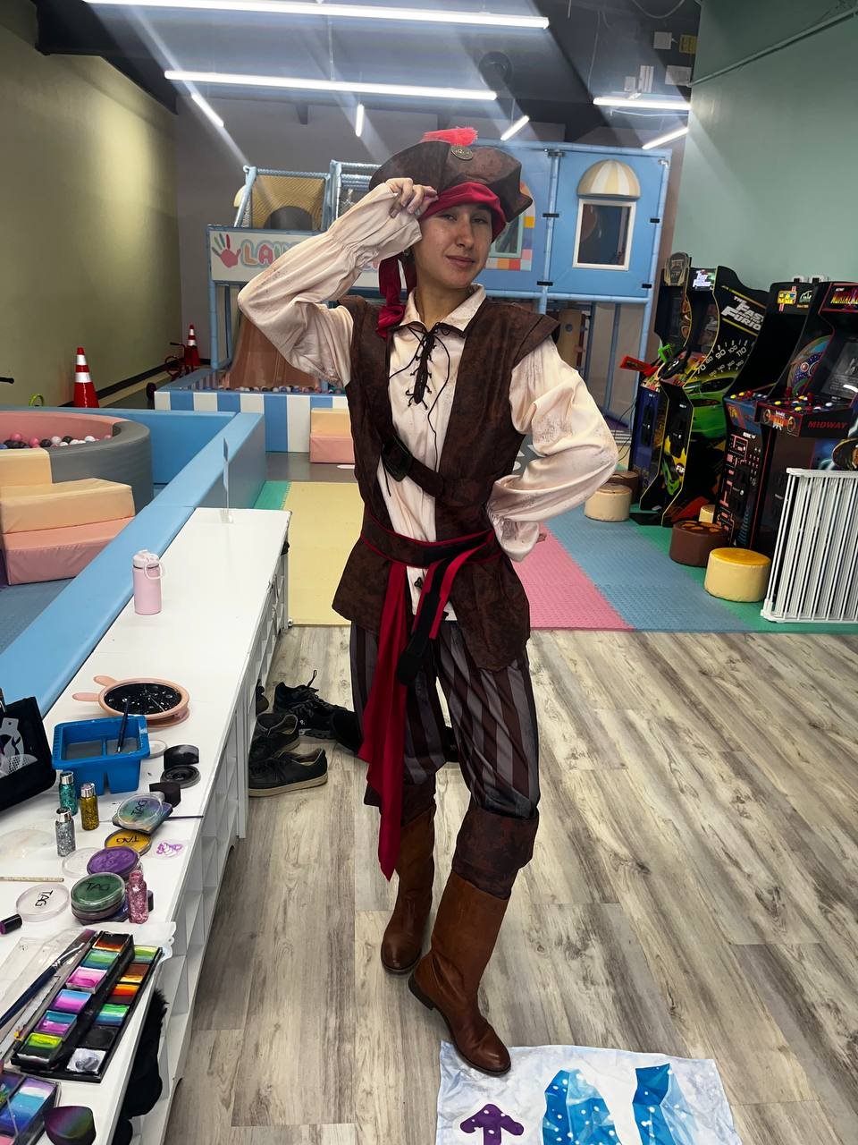 Person dressed as a pirate with a pirate hat, red bandana, brown vest, striped pants, and brown boots posing in an indoor arcade or play area.